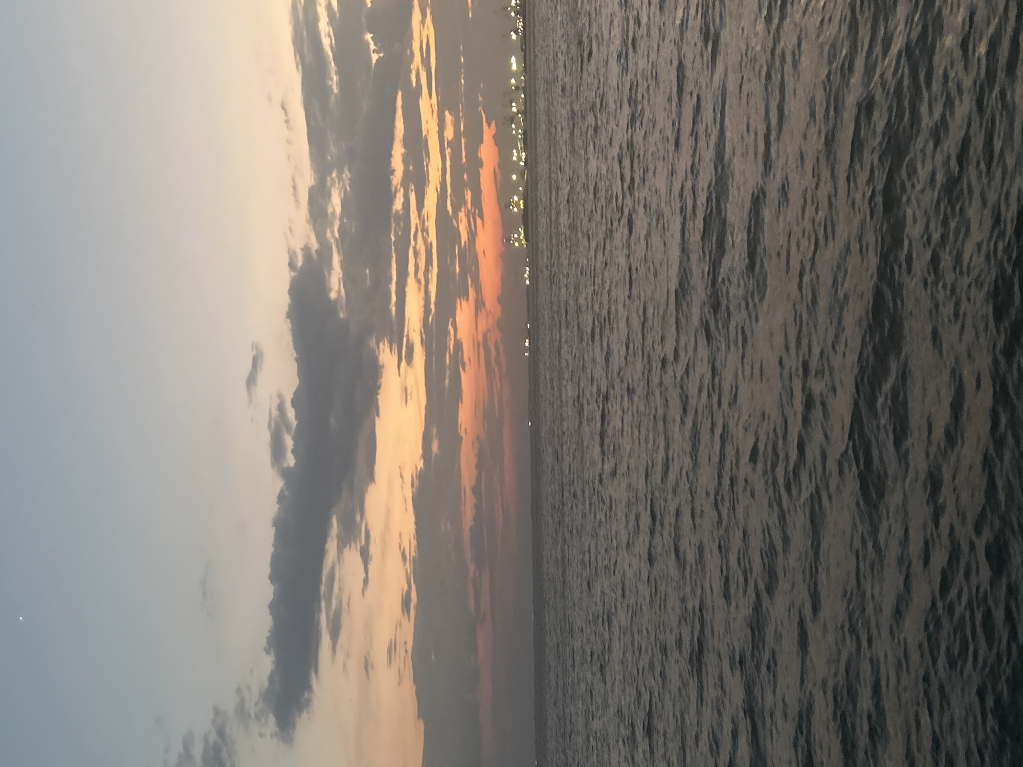 A serene seascape at dusk, likely captured from a boat or the coast, depicts the Thessaloniki Municipal Unit in Greece. The sky transitions from a pale blue at the top to a warm gradient of orange and pink near the horizon, illuminated by scattered clouds. A faint crescent moon is visible in the upper left portion of the sky. In the distance, a city skyline is faintly discernible, marked by a line of lights and what appear to be industrial cranes, suggesting a port area. The sea in the foreground is calm, with gentle ripples reflecting the twilight colors. No people or specific activities are clearly identifiable.