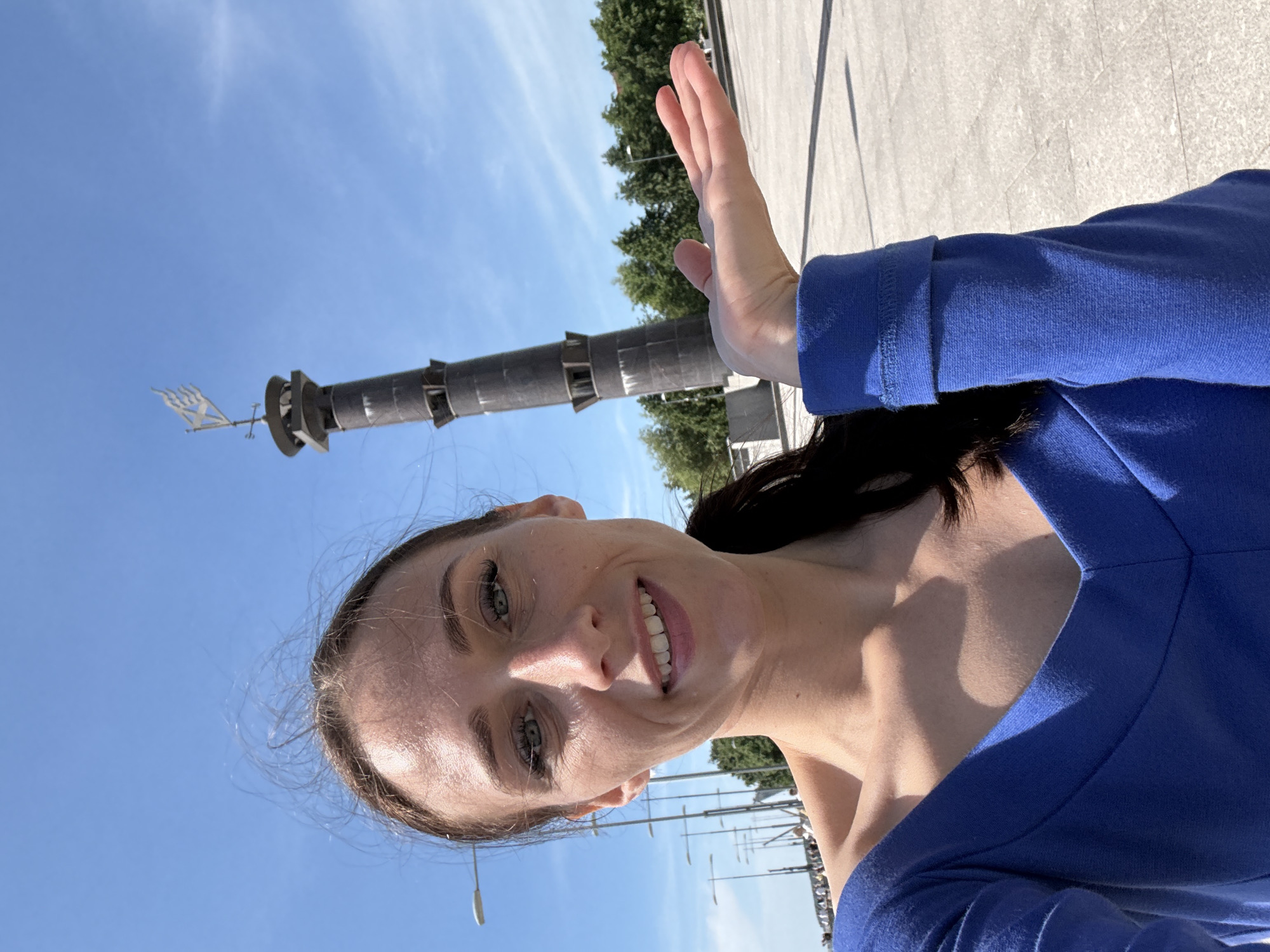 A woman is taking a selfie outdoors in front of a tall, dark, cylindrical monument topped with a weather vane. The monument appears to be made of metal sections with visible openings or platforms. The woman has light skin, dark hair pulled back, and is wearing a blue off-the-shoulder top. She is smiling and holding her right hand up with her palm facing outward, as if presenting something. The background consists of a clear blue sky, a paved walkway, trees, and streetlights. In the distance, there is a bridge or elevated structure with people visible on it. This monument is likely the Narva Triumphal Arch Obelisk in Saint Petersburg, Russia, which is located on Ploshchad Starogo, in front of the Narva Triumphal Arch itself.