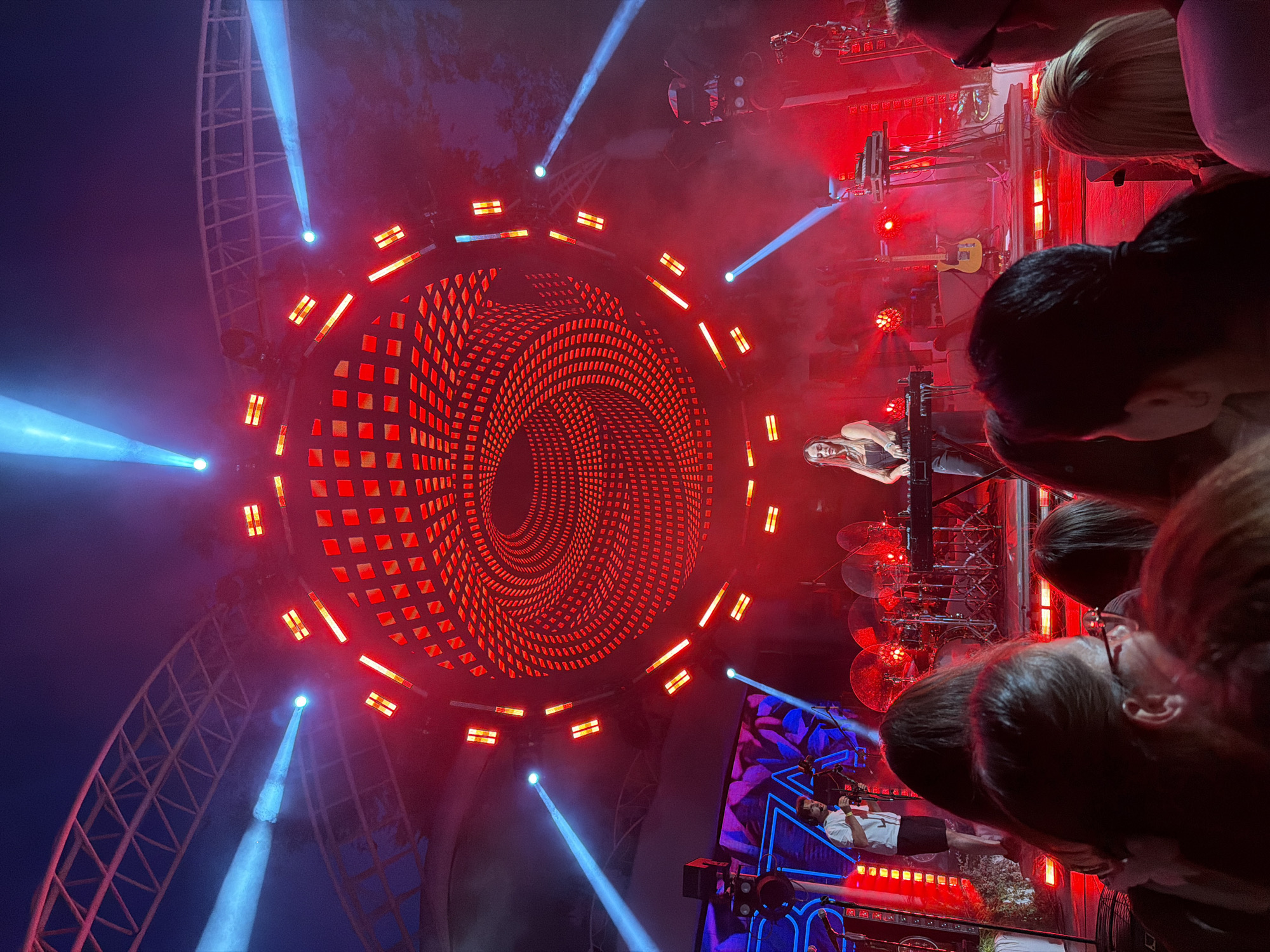 This image captures a live music performance at an outdoor venue, possibly during a festival, in Odesa, Ukraine. The stage is illuminated with a vibrant mix of red and blue lights. A prominent circular LED screen behind the performers displays a mesmerizing, swirling red pattern. Several blue spotlights cut through the smoky atmosphere from above.
On stage, a female DJ is the central figure, standing and operating a DJ console. To her left, a man in a white t-shirt and black shorts is visible, holding what appears to be a camera or microphone on a stand, suggesting he may be a technician or interviewer. Drums and a yellow electric guitar are also visible on the stage, indicating a live band setup.
In the foreground, a crowd of spectators is gathered, their backs mostly to the camera, engrossed in the performance. The lighting creates a dynamic and energetic ambiance. The architectural structure supporting the stage lighting is visible in the background, suggesting a large outdoor setup. The time of day appears to be evening or night, given the intensity of the artificial lights.