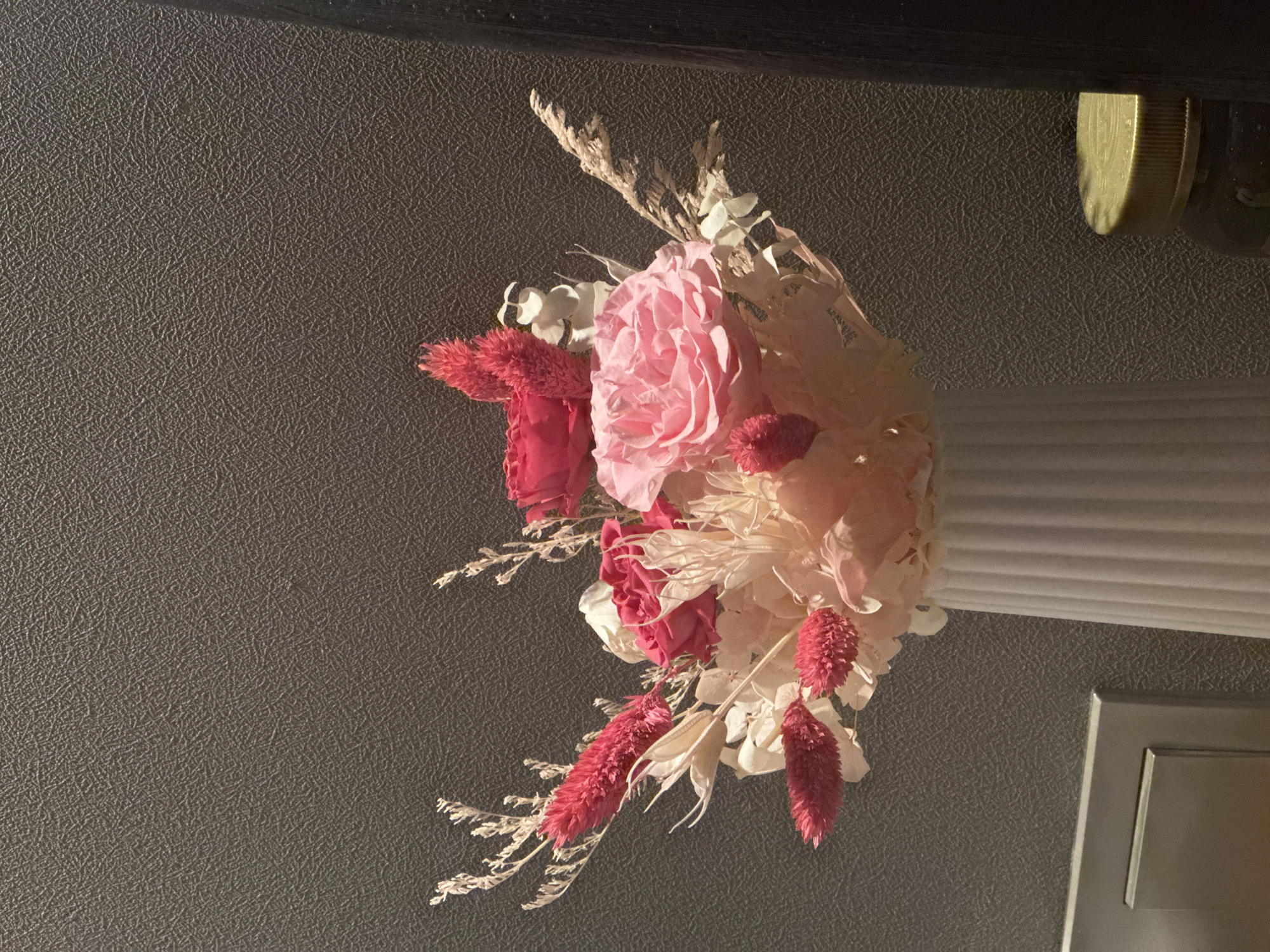 A floral arrangement is suspended from a molding in an interior space. The arrangement includes pink roses and various white and reddish-pink dried flowers and grasses. The wall behind the arrangement is a textured gray. To the right, a white, horizontally-grooved molding is visible, along with a portion of a silver-colored rectangular panel. Above the arrangement is a dark, possibly black, element, possibly a ceiling or another part of the structure. A small, round gold-colored object is also affixed to the structure. The location is Unknown, Unknown.
