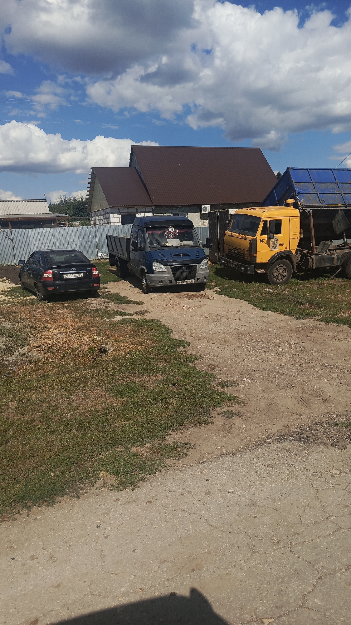In the city of Oktyabrsky Gorodok, Russia, on a bright, sunny day with scattered clouds, a scene unfolds in what appears to be a rural or semi-industrial area. The ground is a mixture of gravel and dry grass. A tall, corrugated metal fence runs along the left side of the image.
Parked on the gravel area are three vehicles. On the far left is a black sedan, likely a Lada Granta, with the license plate X884OX 86. To its right is a blue Gazelle van, facing forward, with its bed empty. A sticker of a cartoon character is visible on the front windshield. The license plate of the Gazelle reads B157BH 36. Next to the Gazelle, to its right and slightly further back, is a yellow Kamaz truck, its cargo bed raised. The truck is weathered and shows signs of use.
In the background, several buildings are visible. Two of them have dark brown, corrugated metal roofs. One building on the left has a lighter colored roof and appears to be a garage or workshop. Another building behind the Gazelle van has a light-colored facade and an air conditioning unit mounted on the wall. The overall impression is of a functional, perhaps slightly disused, area.