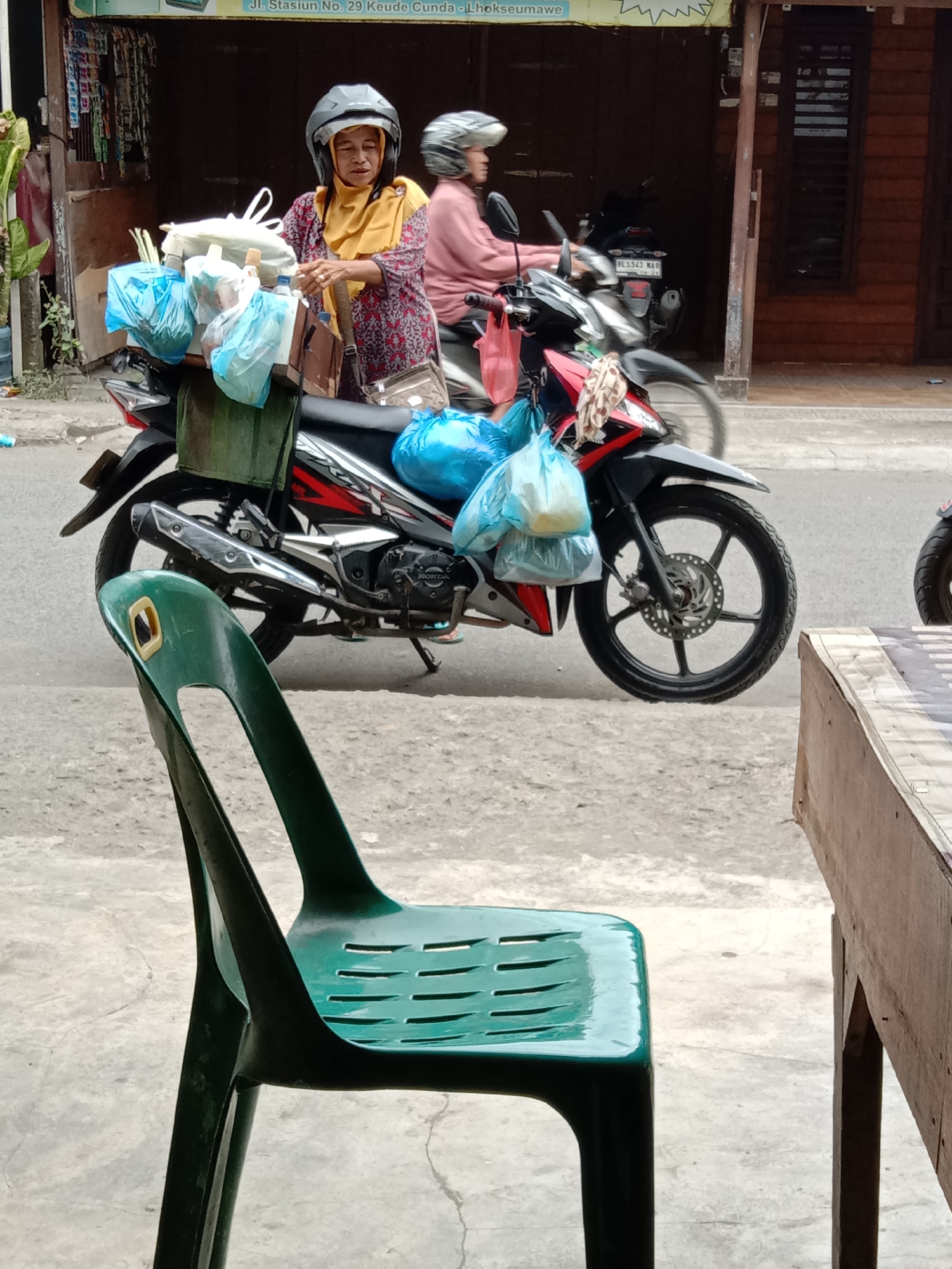 The image captures a street scene in Lhokseumawe, Indonesia. In the foreground, a green plastic chair is prominently featured, partially obscuring the view. Behind the chair, a person wearing a yellow hijab and a patterned shirt is seated on a motorcycle, appearing to be engaged in a transaction or conversation. They are wearing a helmet. The motorcycle is loaded with several blue plastic bags and other items. Another person, also wearing a helmet, is seated on a motorcycle behind the first one, their face obscured by the helmet. The background shows the exterior of buildings with wooden elements. The ground is paved with concrete. The overall impression is a snapshot of daily life on a street in the city.