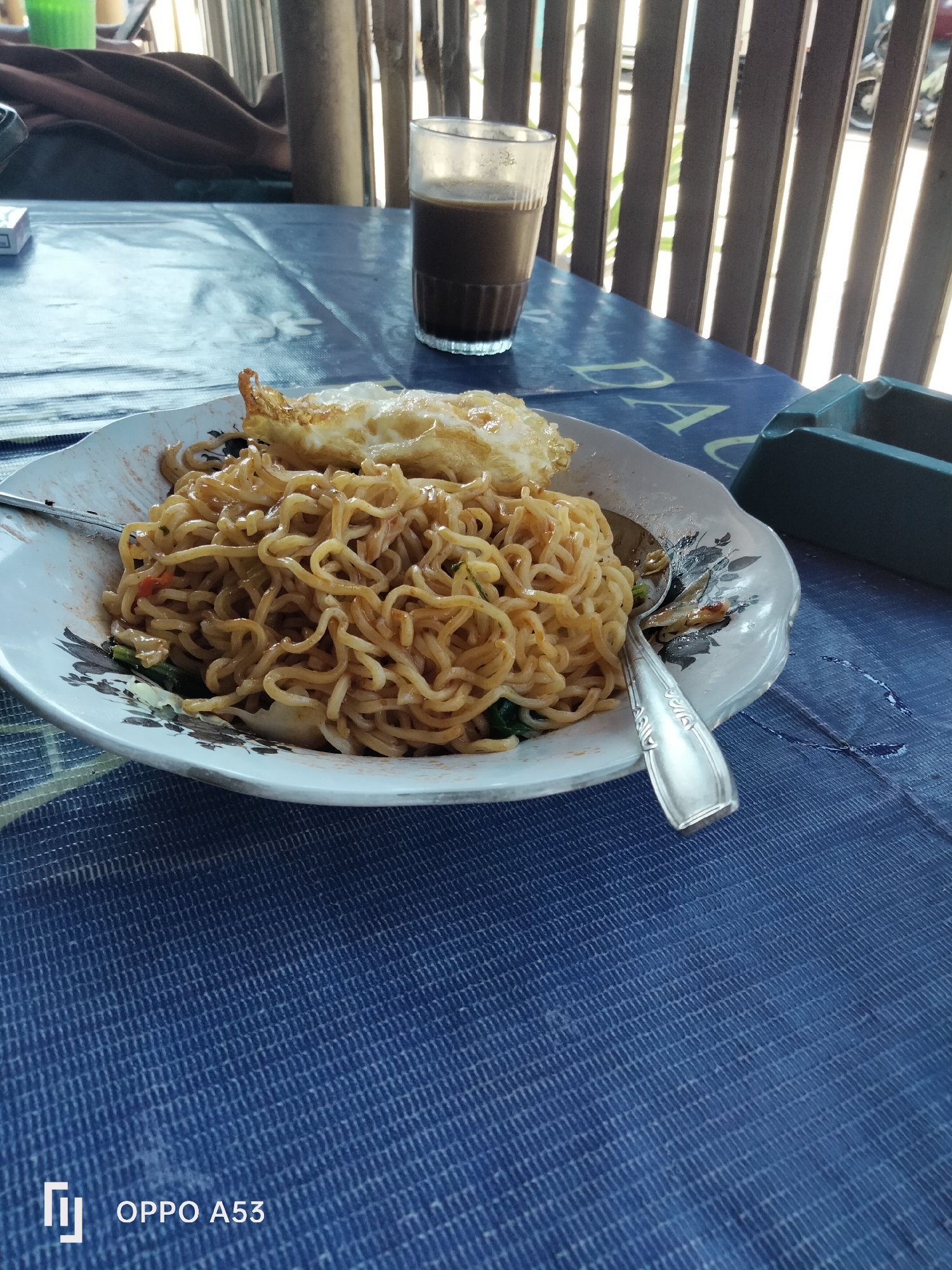 A plate of Indonesian fried noodles, known as Mie Goreng, is presented on a blue tablecloth at an outdoor food stall in Denpasar, Indonesia. The dish features a generous portion of noodles, topped with a fried egg. A small glass of dark liquid, likely coffee or tea, sits to the right of the plate. An ashtray is visible further to the right, suggesting a casual dining environment. The background shows a wooden fence with vertical slats, behind which sunlight filters through, indicating an open-air setting. The presence of a cigarette pack on the left side of the image reinforces the impression of a typical roadside eatery. The tablecloth has a subtle pattern and the plate has a decorative floral rim. The overall scene captures a moment of everyday life and a popular local meal in Denpasar.