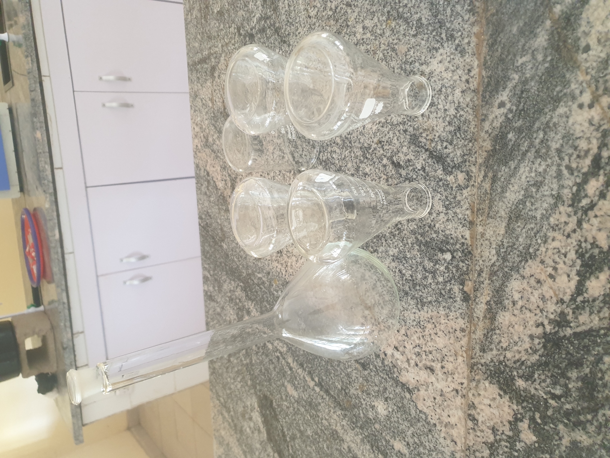 The image shows a collection of laboratory glassware arranged on a speckled granite countertop. In the foreground and to the left, a clear glass volumetric flask with a narrow neck and a ground glass stopper is prominently displayed. To the right of the flask, four clear glass beakers of varying sizes are visible. Two of the beakers appear to be funnel-shaped with graduated markings, one with "250ml APPROX" and the other with "200ml APPROX". The other two beakers are cylindrical, one also marked "200ml APPROX". In the background, a white cabinet with silver handles is visible, and above it, a laboratory bench with some equipment, including a red and white plastic dish. The scene appears to be set in a laboratory, possibly in Jalingo, Nigeria, given the mention of the city. The lighting suggests an indoor setting. There are no people present in the image.