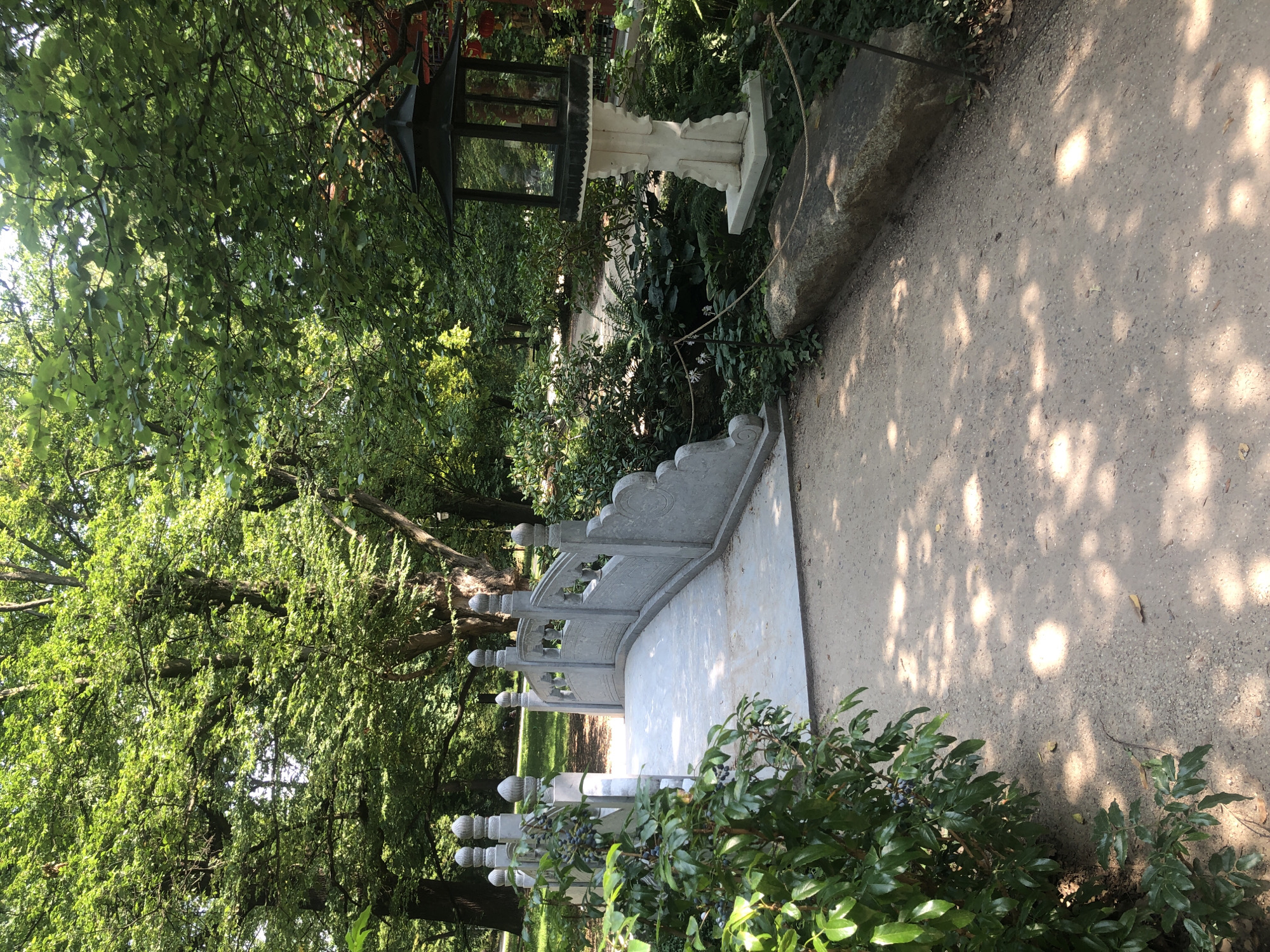 This is a photograph of a stone bridge in a park setting. The bridge is gray and arched, with decorative balustrades topped with finials. It spans across a path covered with gravel. To the left of the bridge, there is a lush green bush with dark berries. To the right of the path, there is a stone lantern on a pedestal, surrounded by dense greenery. A rough-hewn stone slab serves as a bench or border on the right side of the path, with a rope demarcating the edge. The background is filled with mature trees with dense green foliage, casting dappled shadows on the path. The overall impression is of a tranquil, shaded garden.
This scene is located in the Japanese Garden within the Łazienki Park in Warsaw, Poland.