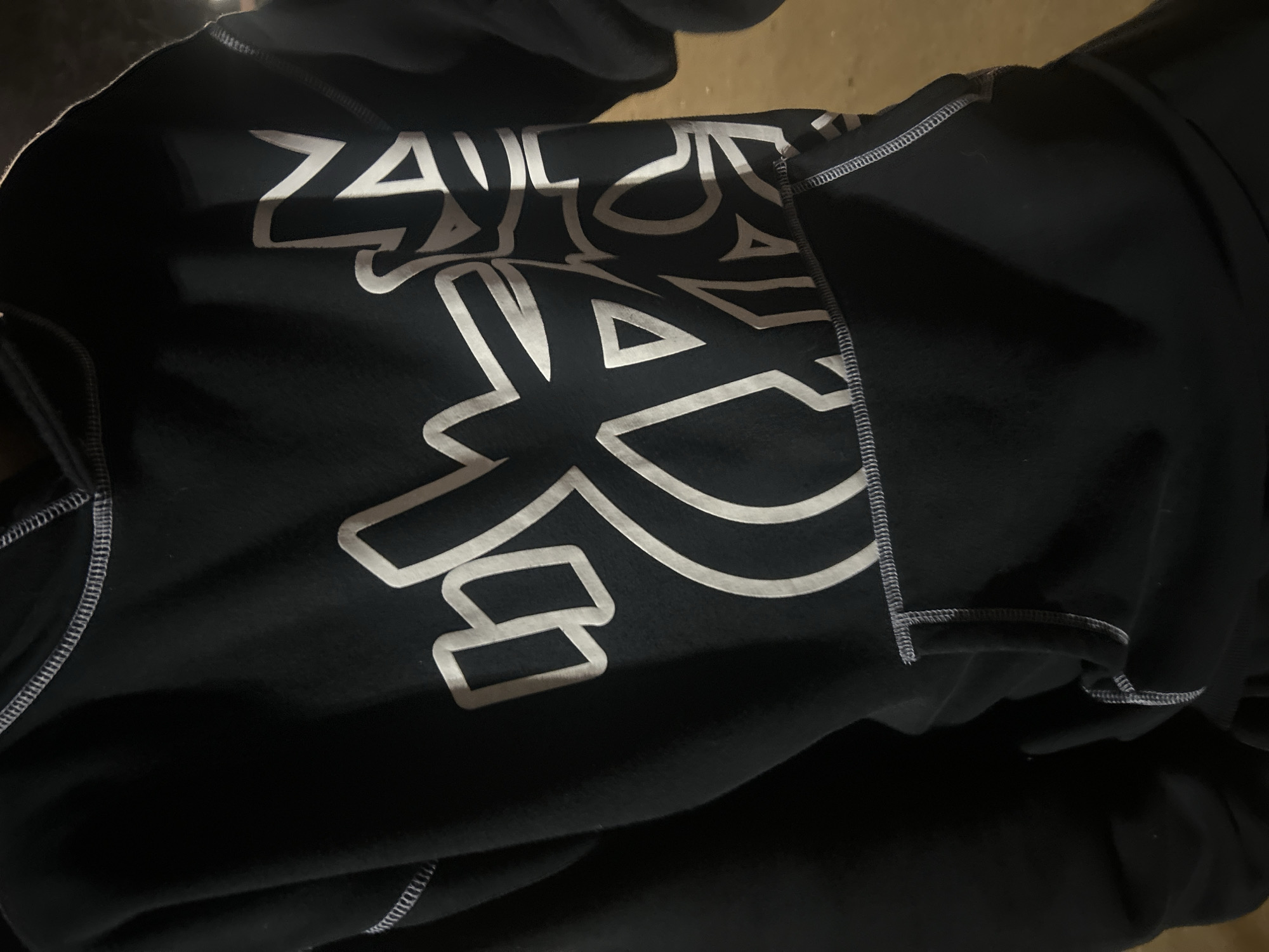 The image shows a black hoodie with a white graphic design. The design appears to be a stylized logo or lettering. The hoodie has visible stitching details along the seams. The background is out of focus and dimly lit, suggesting an indoor environment. There is no indication of any location in Pazardzhik, Bulgaria. The image only contains a piece of clothing.
