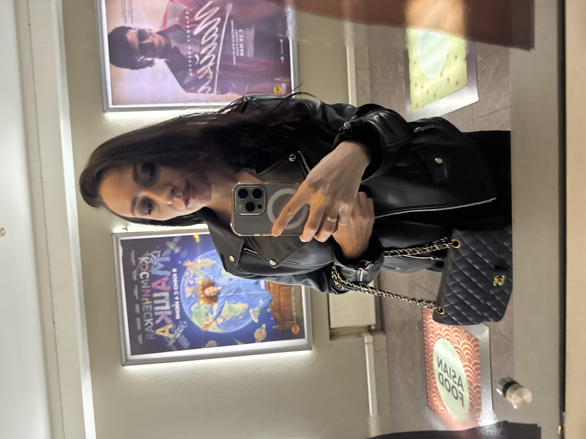 A woman in a black leather jacket takes a mirror selfie in Saint Petersburg, Russia. Behind her are posters. To the right, a sign reads "Asian Food". She is holding a phone with an Apple logo. She is wearing a black shoulder bag.
