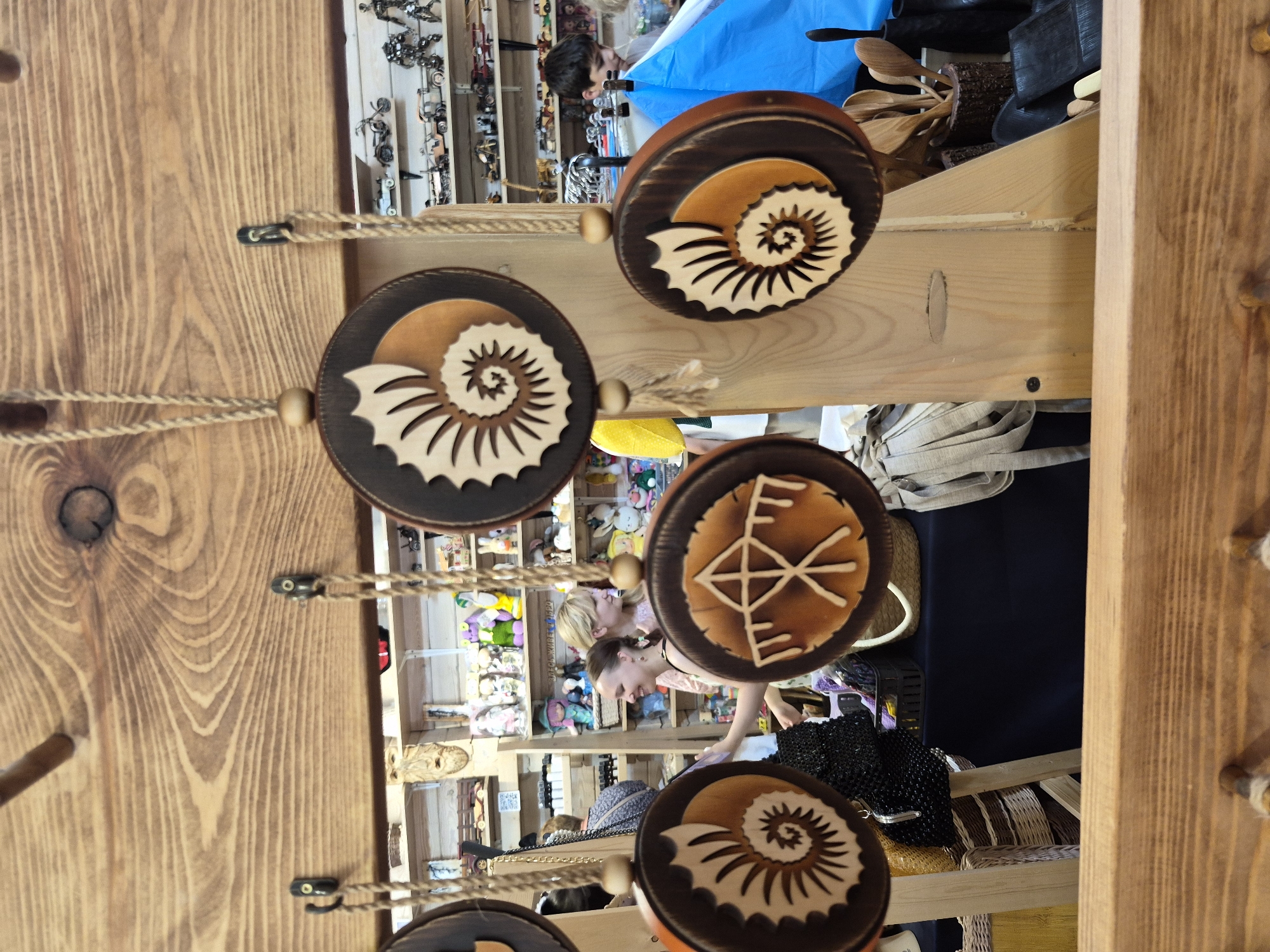 The image depicts an indoor market stall with wooden shelves displaying various items. In the foreground, several circular wooden ornaments with carved ammonite designs are hanging from ropes. Behind the ornaments, a man and two women are visible. The women appear to be browsing the stall, and one is interacting with a display of goods. The shelves behind them are filled with a variety of objects, including what appear to be small toys and decorative items. The overall impression is of a lively marketplace. The city of Krasnyy Klyuch, Russia, is mentioned as the location, suggesting a cultural context for the market and its wares.