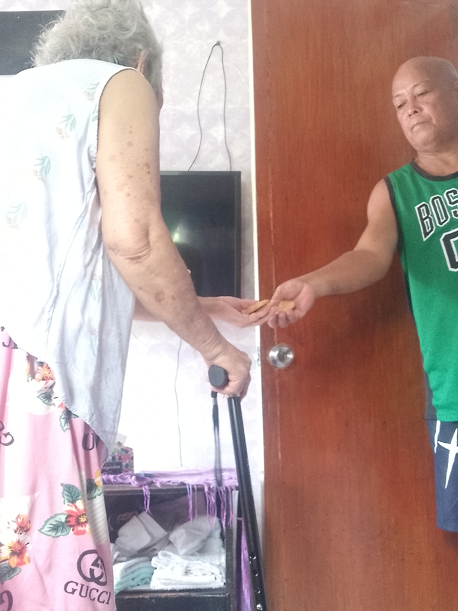 Inside a residence in Davao City, Philippines, an older adult female with gray hair, wearing a white floral top and pink floral Gucci bottoms, holds a black cane in her left hand. An adult male with a shaved head, wearing a green basketball jersey with "BOS" visible, extends his right hand holding several small, round, light brown objects to the female. A brown door is behind the man. A small cabinet with white towels and a purple scarf is partially visible near the female. A television and a thin, white cord are on the wall behind the cabinet.