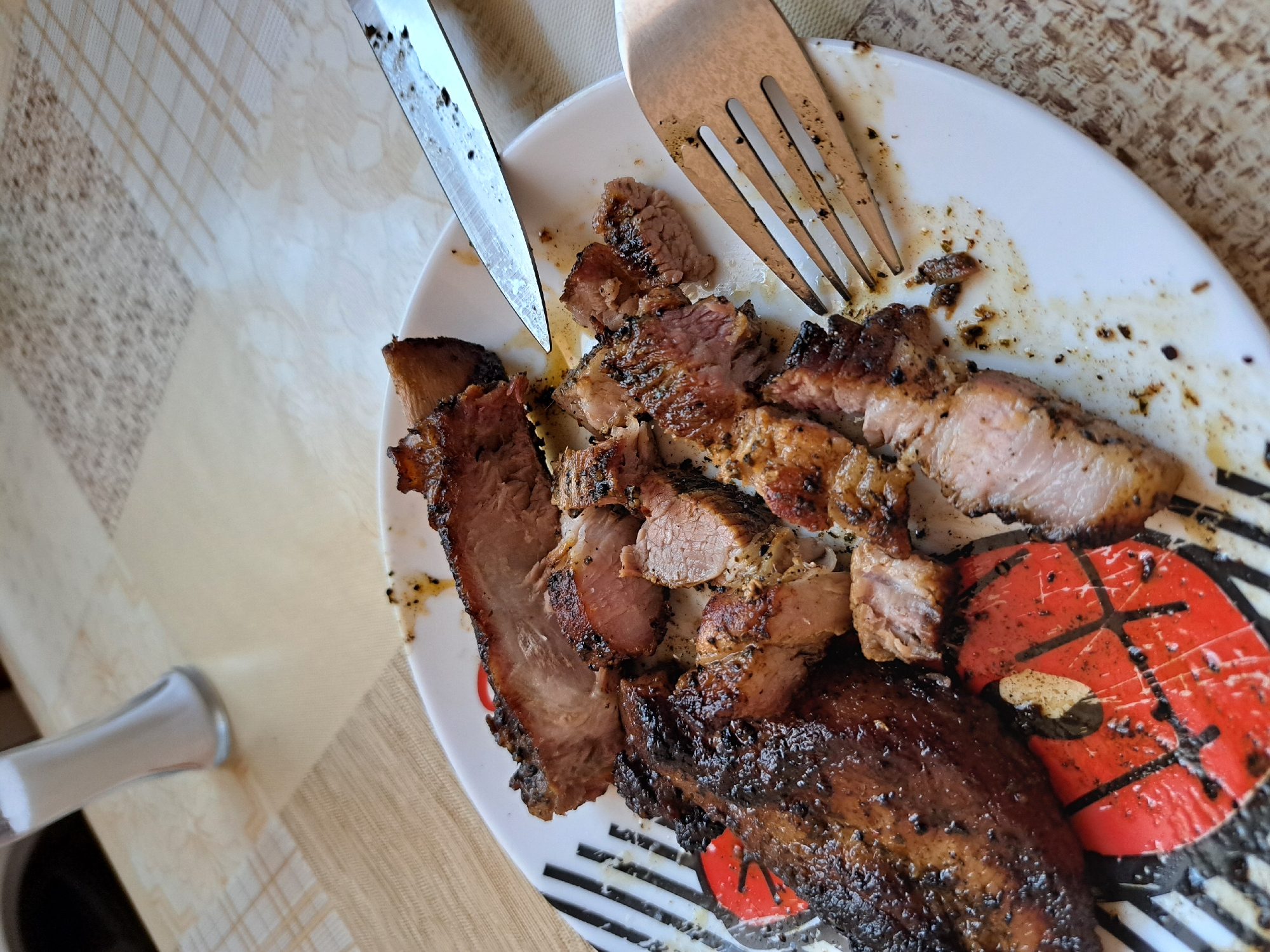 A close-up shot shows a plate of cut grilled meat, a fork, and a knife on a patterned tablecloth. The meat appears to be pork, with a browned exterior and a pinkish interior. The plate has a cartoon design featuring black stripes and red accents. The scene is indoors, but the specific location within the city of Unknown, Unknown cannot be determined from the image.