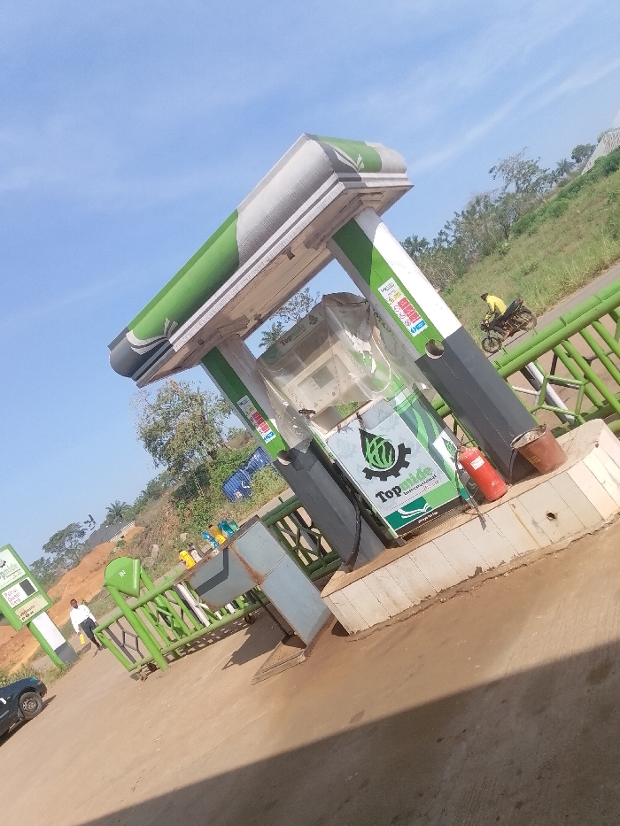 This image captures a gas station operating under the brand "TopWide International" in Ayegbaju, Nigeria, under a clear blue sky. The central structure consists of a green and white canopy supported by pillars. Beneath the canopy, two green and white fuel pumps are visible, one of which is partially encased in clear plastic sheeting. A red fire extinguisher and two rusty metal buckets rest on the concrete base adjacent to the foreground pump. A green metal fence separates the immediate station area from a paved road. Beyond the road, a steep, grass-covered embankment with scattered trees rises. On the left side of the frame, a dark-colored vehicle is partially visible, and a man in a white shirt and dark trousers stands near a "TopWide International" sign. In the background, a person wearing a yellow shirt is operating a motorcycle on the road, traveling from right to left.