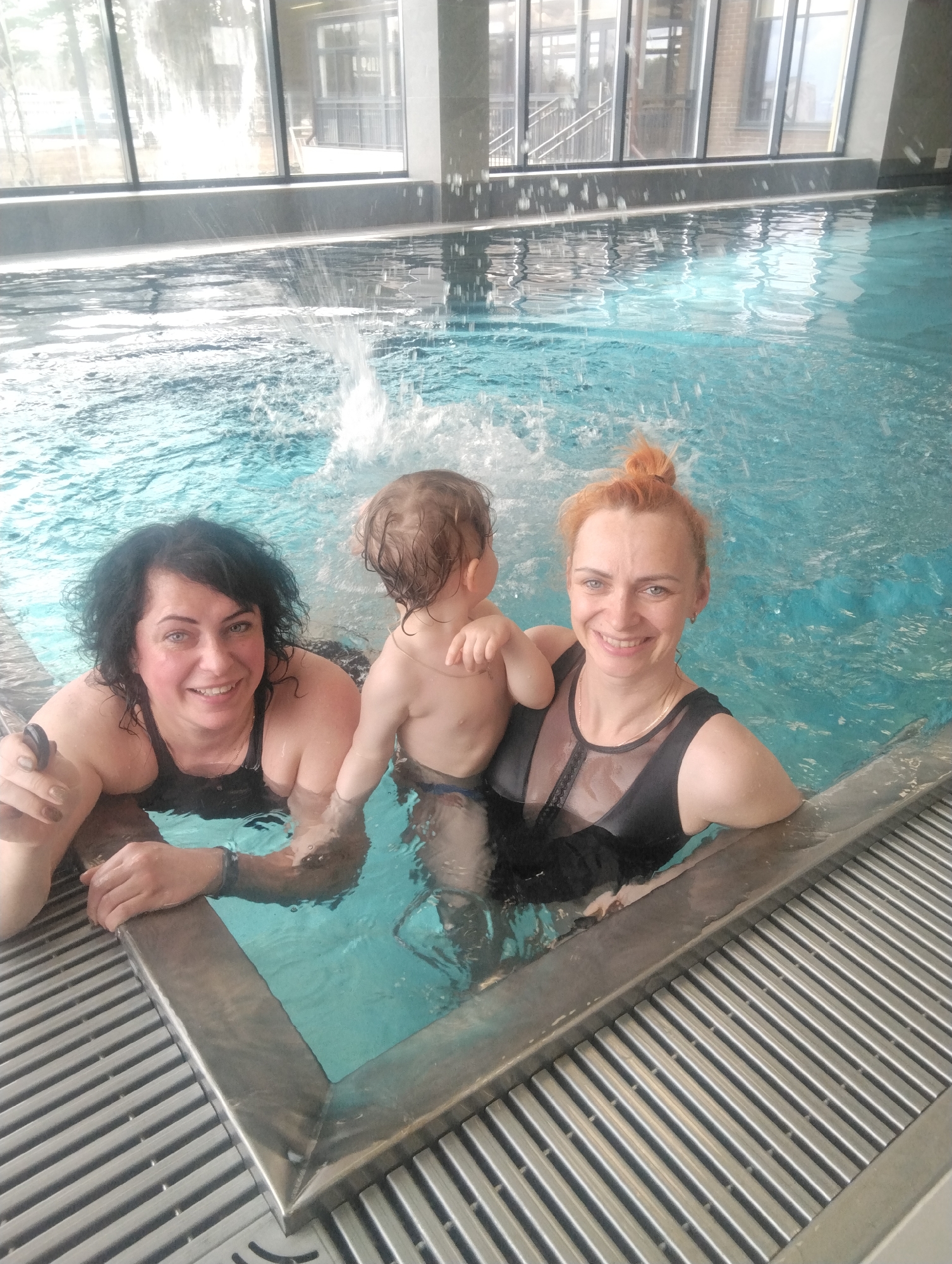 This media file captures a joyful scene within an indoor swimming pool in Chaikovsky, Russia. The main subjects are two adult women and a baby or toddler, all partially submerged in the bright blue water.
The woman on the left has dark, curly hair and is wearing a black swimsuit. She is smiling directly at the camera, her left arm resting on the pool's edge, and appears to be holding a dark wristband or small device. In the center, a baby with light brown hair is seen from behind, facing away from the camera, standing in the water with splashes erupting around them. The woman on the right, sporting reddish hair tied in a bun, is also smiling at the camera. She wears a black swimsuit with sheer, possibly lace, detailing on the chest, and is leaning against the pool edge.
The background features large, floor-to-ceiling windows that let in abundant daylight, suggesting it is daytime. Through these windows, glimpses of a modern building with architectural details like window frames and what appears to be an exterior staircase are visible. The pool itself has a clean, contemporary design, with metallic or grey slatted grates bordering the edge, likely part of an overflow or drainage system. Water is visibly active, with splashes near the center where the baby is. Both women exhibit happy and relaxed expressions, indicating a pleasant experience. There is no discernible text visible in the image.