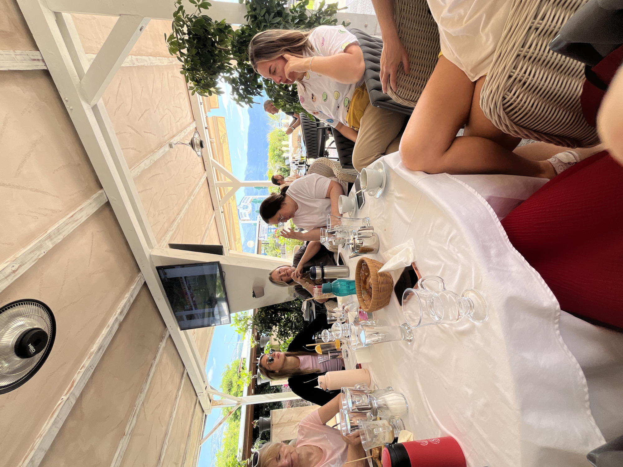 The image shows a group of people at a table in an outdoor restaurant or cafe in Budva, Montenegro. They are sitting around a large, round table covered with a white tablecloth, cluttered with glasses, cups, bottles, and other dining items. Several individuals are visible; one woman is wearing a t-shirt with smiley faces and khaki-colored pants, while others are wearing sunglasses, t-shirts and other clothing, and are seated at the table. The background includes a view of the Budva coastline with mountains in the distance. Above the table is a roof structure with a TV mounted on one of the supports and a heater. The general setting is relaxed and casual, suggestive of a daytime meal.