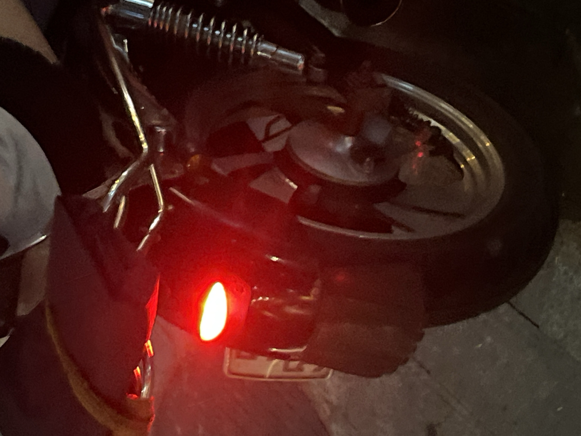 The image shows the rear of a black motorcycle parked on a paved surface, likely in an urban environment. The motorcycle has a bright red taillight illuminated. A license plate is visible below the taillight, appearing to be a white plate with dark lettering. The motorcycle features chrome accents, including a rear shock absorber and part of the wheel rim. The ground appears to be concrete or asphalt, with some faint markings or texture. The lighting is dim, suggesting it may be nighttime or a poorly lit area. The background is dark and out of focus, providing little context about the surrounding environment. There are no people clearly visible in the image.