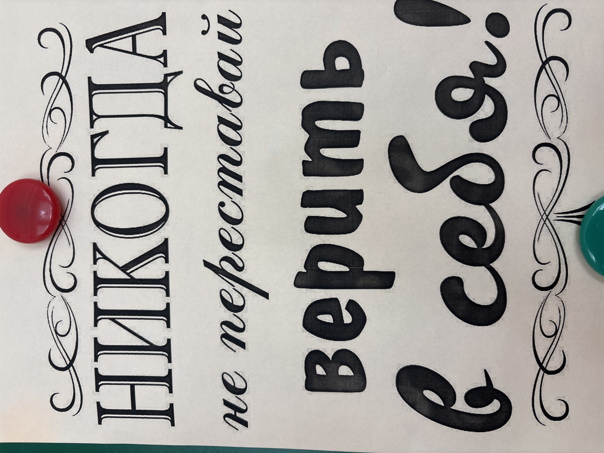 The image is a close-up of a piece of paper with text written in Russian. The text is in black ink and appears to be a motivational quote. The letters are stylized, with some in a serif font and others in a script font. The paper itself is a light cream color and has decorative swirls at the top and bottom. There are two round, colored objects attached to the paper: a red one at the top and a teal one at the bottom. The image does not show any specific location in the city of Korovo, Russia, as it focuses on the text and decorative elements.