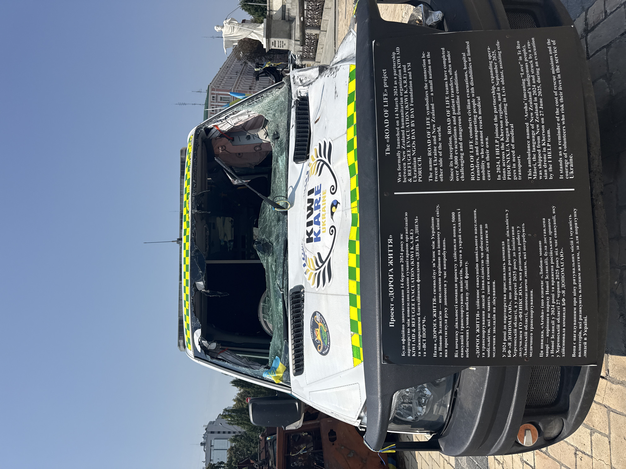 The image shows the front of a damaged white van with a cracked windshield and a checkered yellow and green stripe along the top. The van has logos and text on its side and a black placard in front of it. A Ukrainian flag is visible inside the van's broken windshield. In the background, a white statue of a person with outstretched arms stands in front of a building. To the left of the van, another damaged vehicle, possibly also a vehicle, is visible. The scene appears to be outdoors in an urban environment, suggesting it is located in Kyiv, Ukraine, given the presence of Ukrainian flags and text in Ukrainian. The placard details a project called "The ROAD OF LIFE" and describes its origins and activities, including the evacuation of people and medical transport. The overall impression is one of damage and resilience, possibly related to the ongoing conflict in Ukraine.