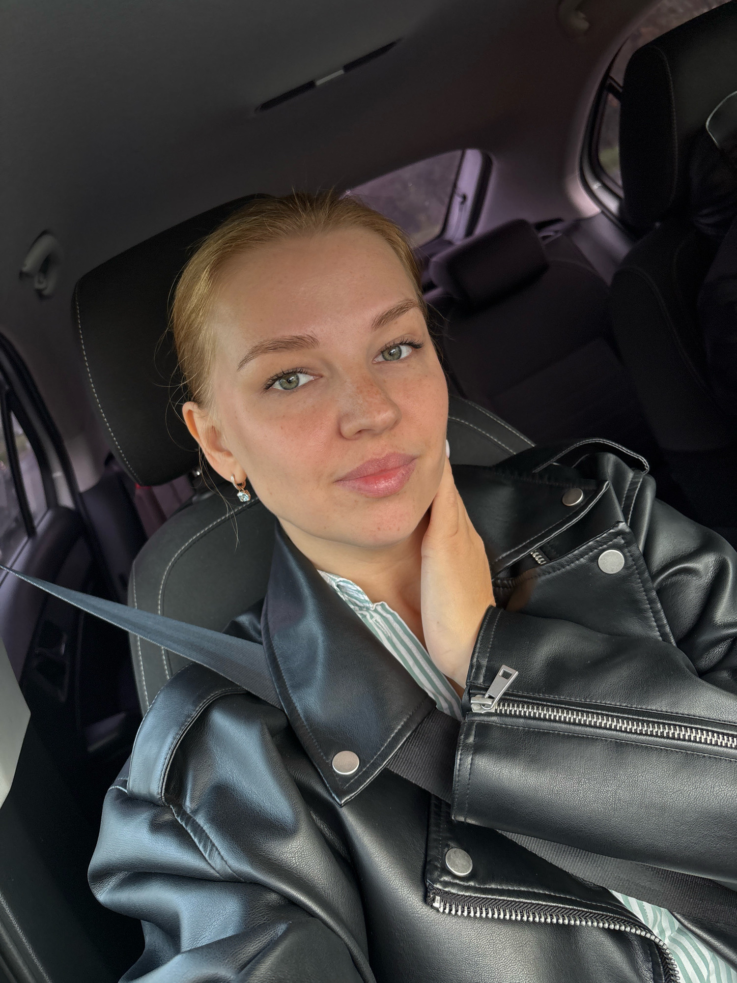 A young woman with blonde hair and green eyes is pictured in the front seat of a car. She is wearing a black leather jacket over a striped shirt and has her hand resting on her neck. Her seatbelt is visible across her chest. The background shows the interior of the car, with dark seats and headrests. The image appears to be a selfie taken by the woman. The location is not specified in the image or metadata.