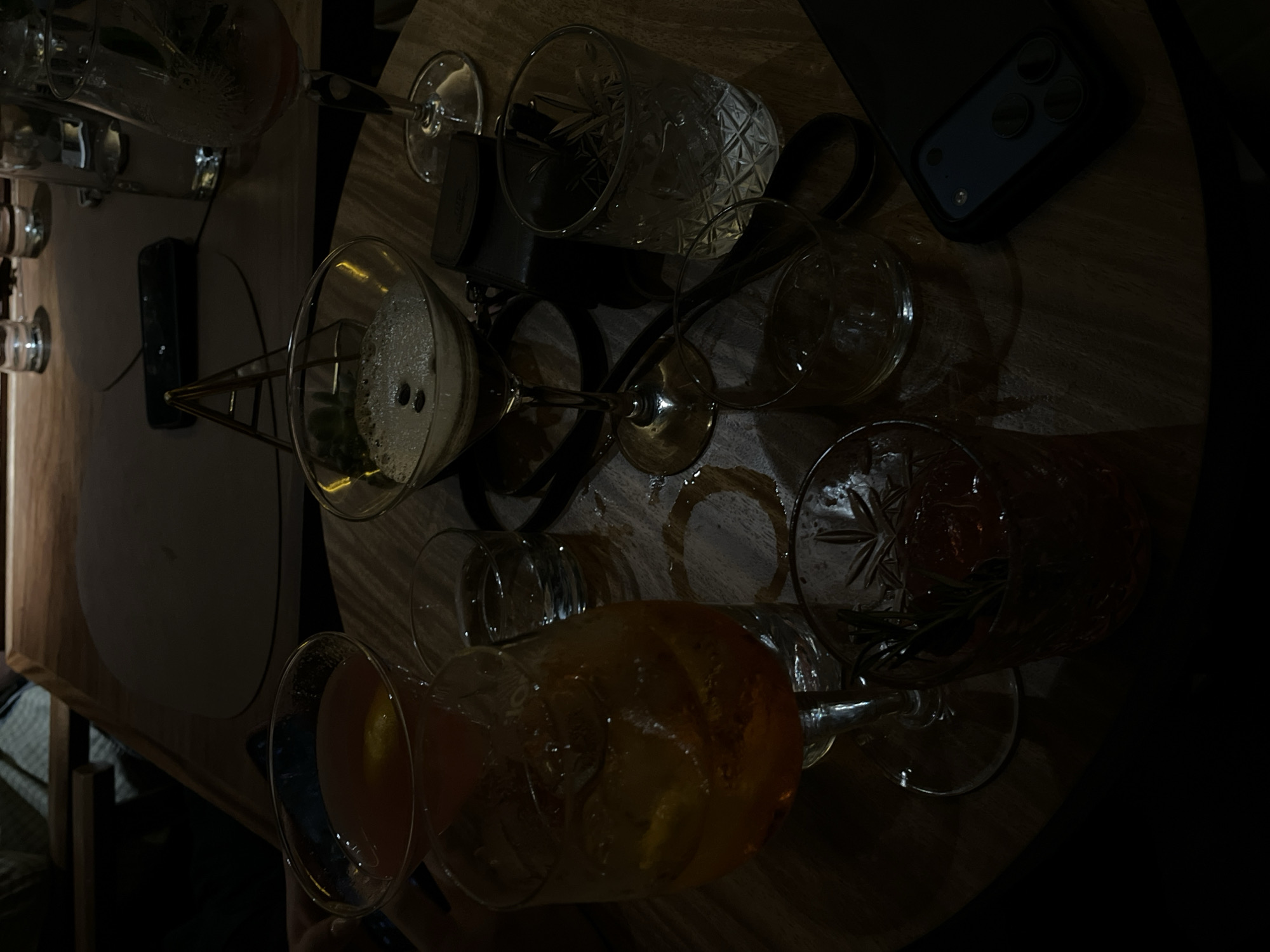 The image depicts a dimly lit, overhead view of a wooden table in what appears to be a bar or restaurant. The table is cluttered with several empty or partially empty glasses and cocktail glasses. There are at least six glasses visible, some with remnants of liquid and ice. A smartphone rests on the table near the upper-right corner of the frame. The environment suggests a social gathering or after-dinner drinks. The specific location in San Andrés Cholula, Mexico is not discernible from the image alone.