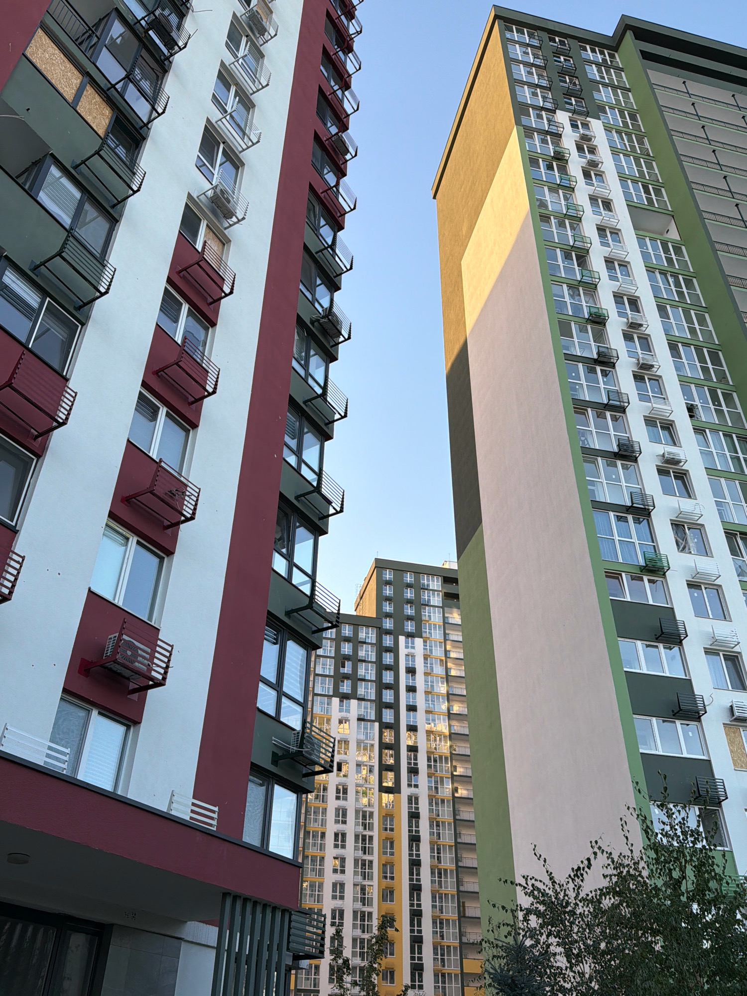 The image depicts a view looking upwards at several modern high-rise apartment buildings in Kyiv, Ukraine. The buildings feature varied architectural designs and color palettes. On the left, a building is characterized by broad vertical stripes of white and dark red, with protruding balconies and air conditioning units visible. In the center and right, a taller building showcases a striking design with a broad, diagonal cream-colored section, transitioning into a section with dark green and white facades. This building also has numerous windows and balconies. Further in the background, another tall residential building with a patterned facade of windows and balconies is visible, reflecting warm light. The sky above is clear and blue. The presence of some boarded-up windows on one of the buildings might suggest recent damage or ongoing construction. Lush green foliage from trees is visible in the foreground and to the right.