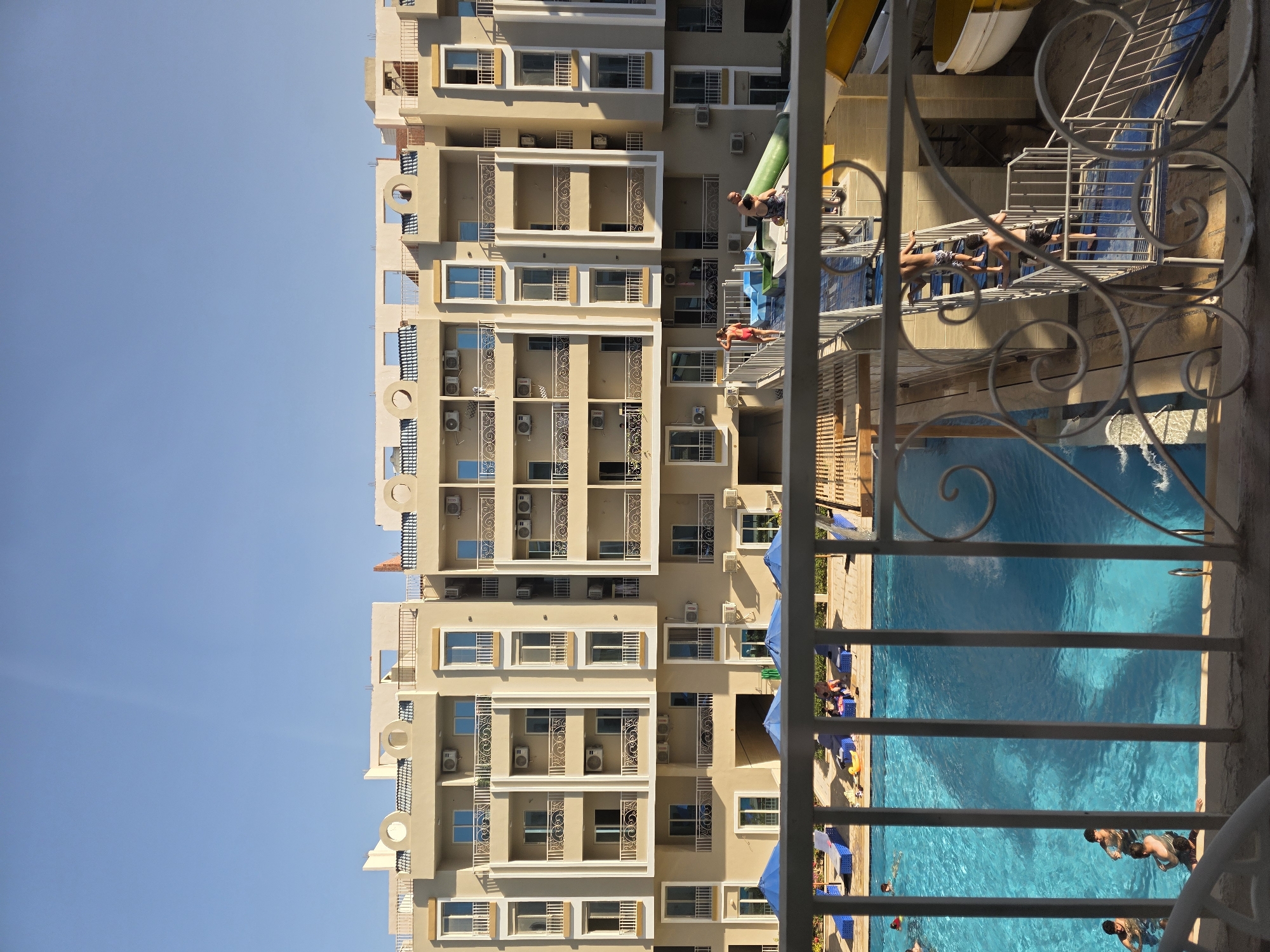 From a high vantage point, possibly a hotel balcony in Hurghada, Egypt, a multi-story building is prominently visible with numerous balconies, windows, and air conditioning units. A swimming pool with people swimming and lounging around it is also visible. The architecture seems consistent with the region. A partial view of a water slide is present. A wrought-iron railing frames the foreground. The sky above is clear.