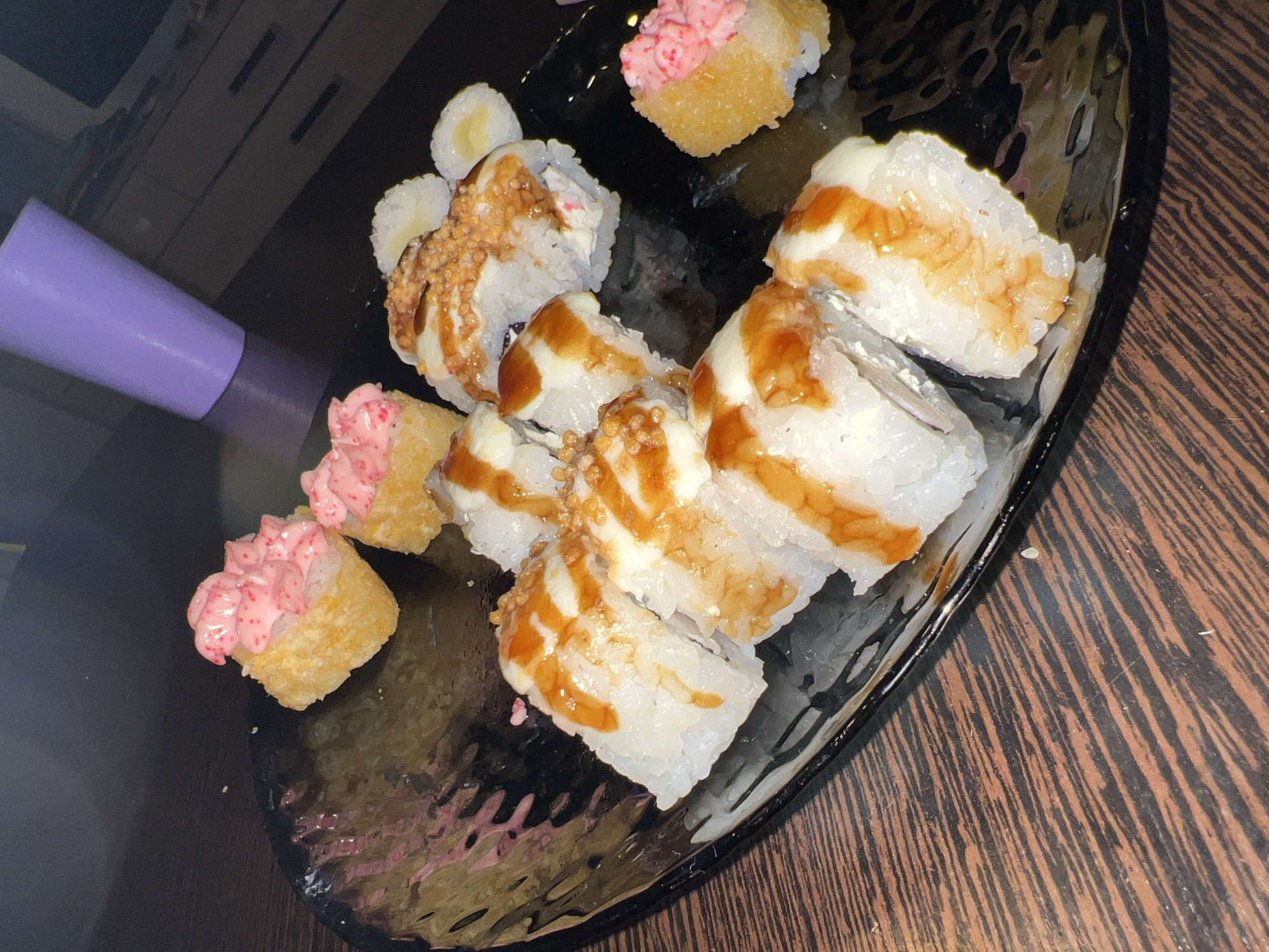On a black plate, sushi is arranged on a wooden surface. Two pieces of sushi are topped with pink sauce. The rest of the sushi is topped with white sauce and a brown drizzle. A purple cylinder is visible on the upper-left side of the frame. The setting appears to be indoors. No people are visible. The location is not discernable.