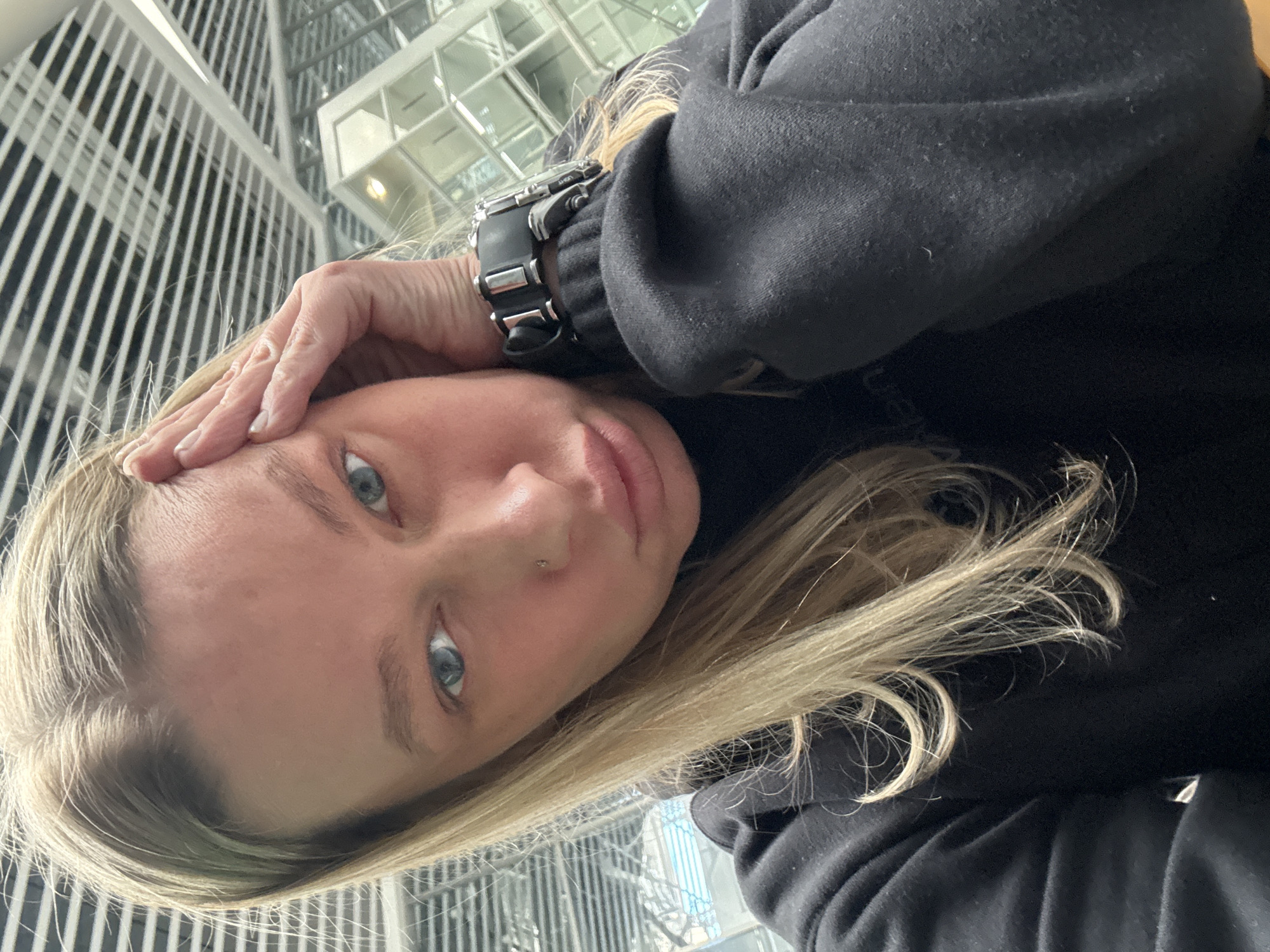 A selfie of a woman with blonde hair and blue eyes. She is wearing a black hooded sweatshirt and a large watch. Her hand is on her forehead. The background appears to be the interior of a modern building with a metal and glass facade, potentially an airport. The location is not Bolshoye Savino, Russia.