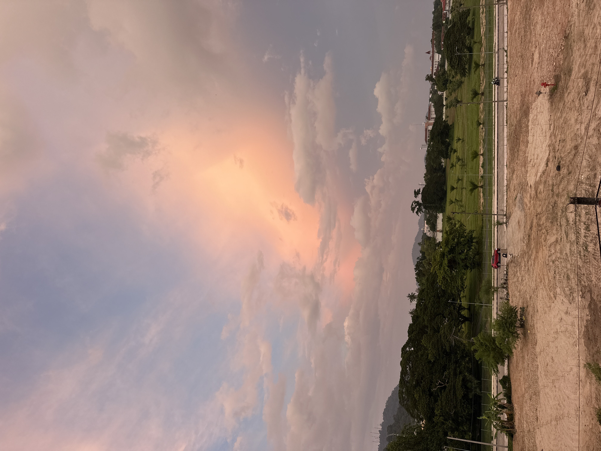 A wide-angle, eye-level, outdoor shot captures a dusky sky over a landscape in Wichit, Thailand. The sky is filled with a dramatic display of clouds, with a large, concentrated patch of vibrant pink and orange light dominating the center, suggesting sunset or sunrise. The lower portion of the image reveals a foreground of sandy, disturbed earth, likely a construction site, marked with small stakes. Beyond this, a paved road runs horizontally, with a red car visible driving on it. Further back, a line of lush green trees forms a dense backdrop, with smaller palm trees interspersed in a manicured grassy field. In the distance, several buildings with red-tiled roofs are visible, nestled among the foliage, and a hazy mountain range forms the farthest horizon. The overall atmosphere is serene and picturesque, with the vibrant sky providing a striking contrast to the earthly elements.
