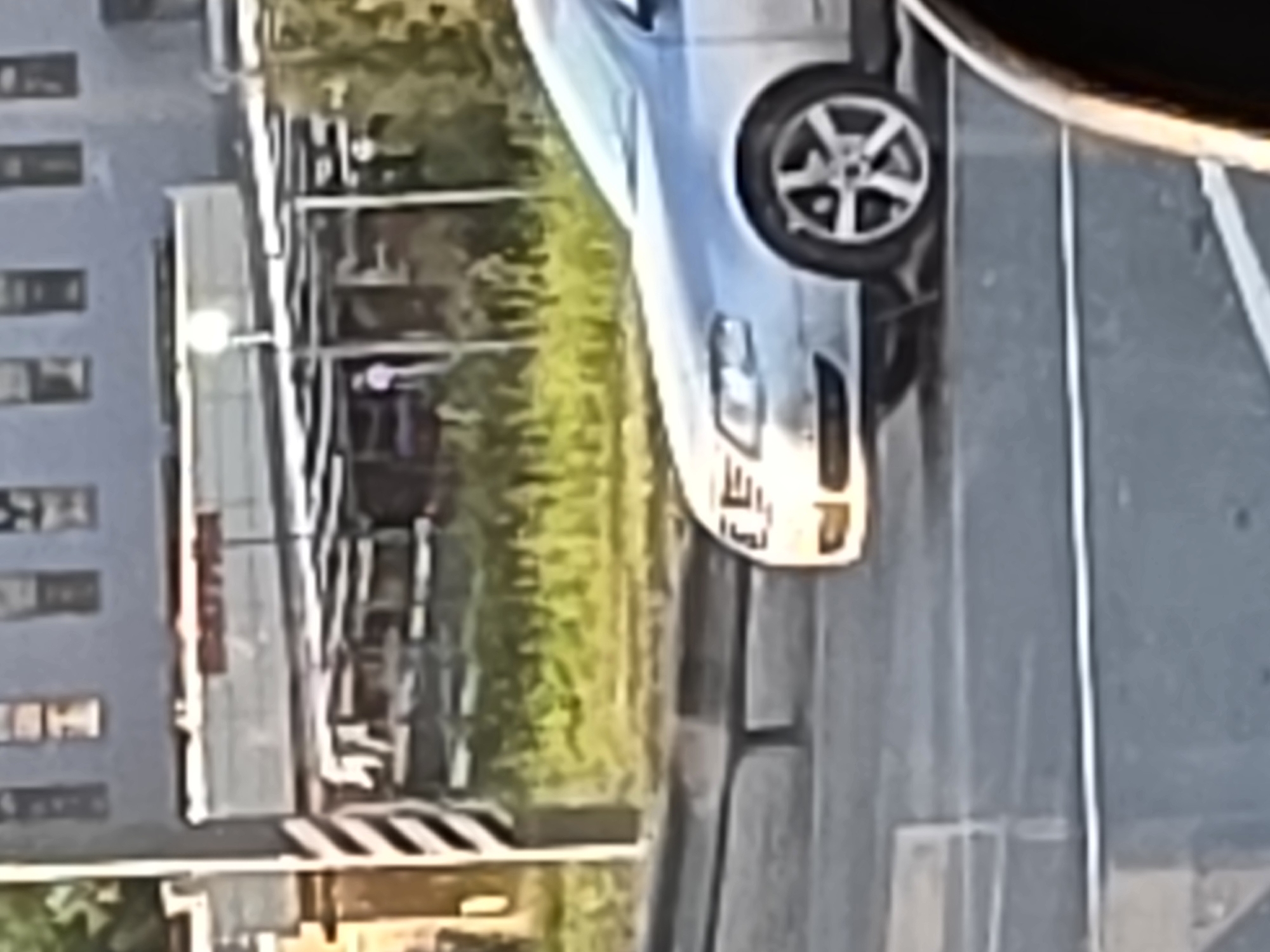 A silver car is parked on the right side of the image, facing left. The car is positioned on a paved surface, with a curb separating it from a grassy area behind. In the background, there is a building with multiple windows and a sign that reads "BROKEN". A streetlamp with a visible light bulb is also present. A person wearing a light-colored garment is standing in front of the building. The overall scene appears to be an outdoor urban environment.