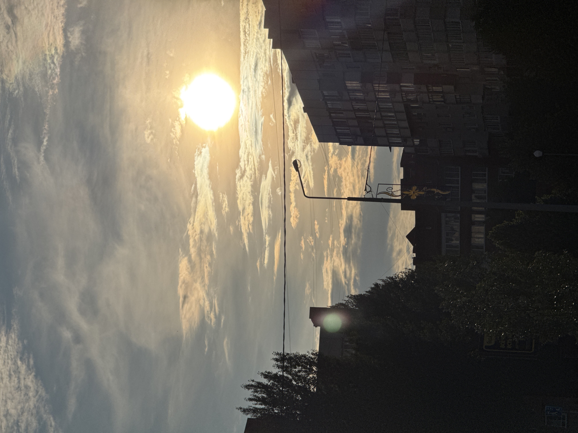 The image captures a sunlit sky above a city. The sun, a bright orb, is positioned in the upper right quadrant of the frame, casting a warm glow on the scattered clouds. The sky transitions from a bright, hazy yellow near the sun to a pale blue at the top. Below the sky, the silhouettes of buildings and trees create a stark contrast.
On the left, tall, dark trees dominate the foreground, their branches reaching upwards. Behind them, a building with visible windows is partially obscured. In the middle ground, a street lamp stands tall, its light fixture a dark silhouette against the sky. To its right, a large, multi-story apartment building is visible, its facade characterized by numerous windows. Further to the right, another building with a stepped roofline is apparent. Power lines are strung across the sky, intersecting the view of the buildings and trees.
The scene appears to be an urban environment, likely a street or residential area, observed during daylight hours with the sun high in the sky. The time of day is suggested by the angle of the sun and the lighting conditions. There are no people or animals visible in the image, and no specific actions or interactions are depicted. The overall impression is one of a quiet, overcast day with intermittent sunlight.