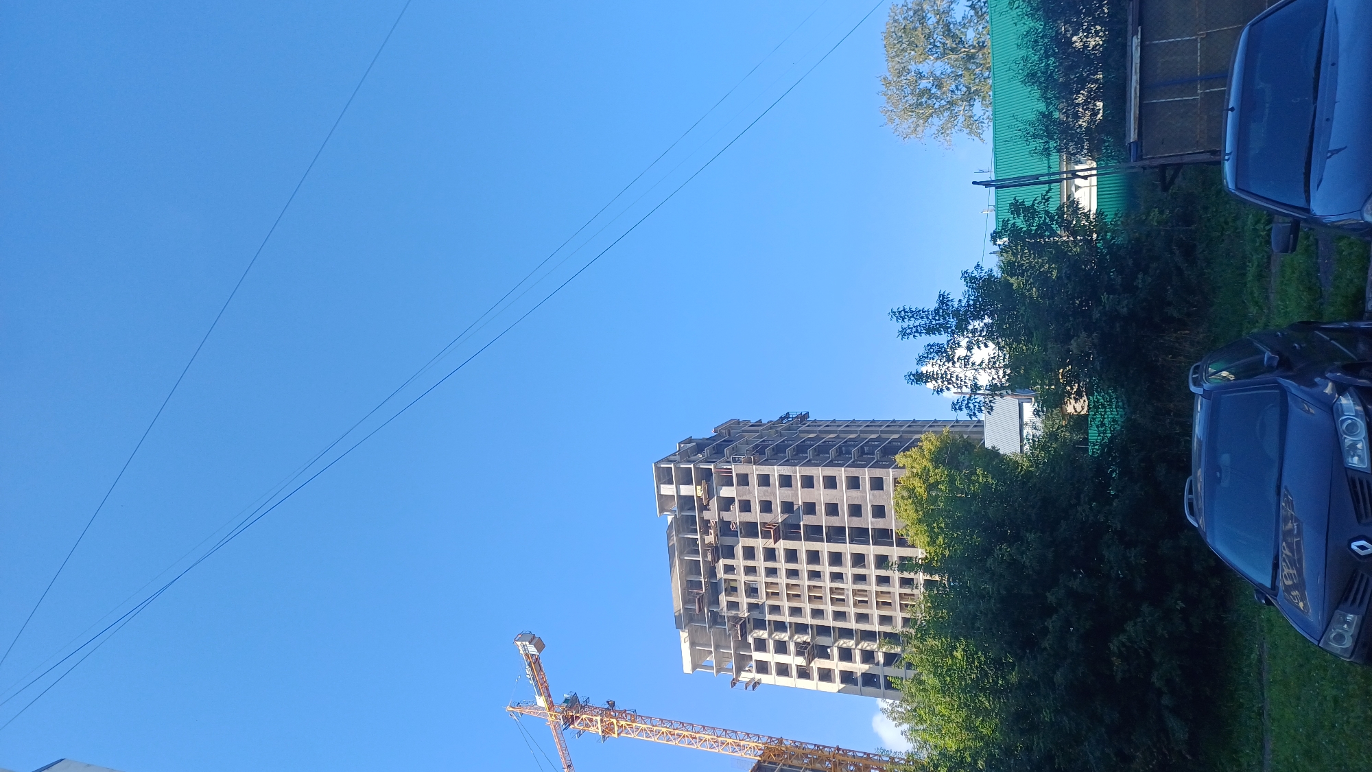 The image shows a construction site with a crane and a building under construction. There are trees and two cars in the foreground. The sky is clear and blue. The image appears to be taken in a city in Izhevsk, Russia.
