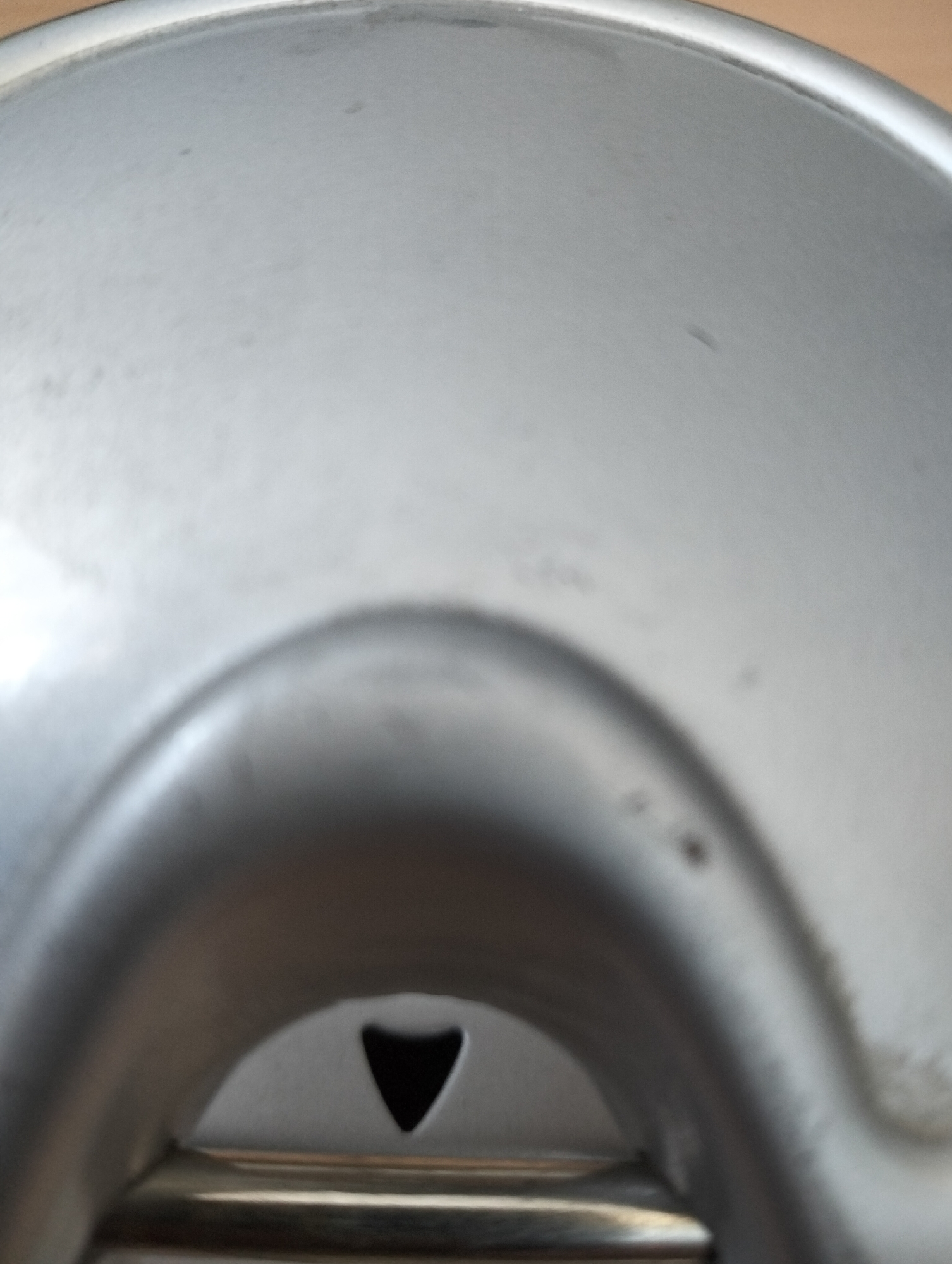 The image shows a close-up, low-angle shot of a metallic surface, possibly the lid or base of a container. The material is silver-toned and appears to be brushed metal, with subtle reflections indicating a light source from above and slightly to the left.
Dominating the lower center of the frame is a concave indentation with a dark, triangular opening or cut-out within it. This opening is deep and appears to lead into a darker space. The metallic surface surrounding this opening is curved and smooth, forming a raised lip or rim. Below this, a horizontal metallic band is visible, with a brushed texture that contrasts slightly with the main surface.
The overall environment is not discernible beyond the immediate object. The lighting is diffuse, causing soft shadows and highlights. The image does not contain any people, animals, or identifiable landmarks. The context of Qarabulaq, Kazakhstan, is not visually represented in the image itself.