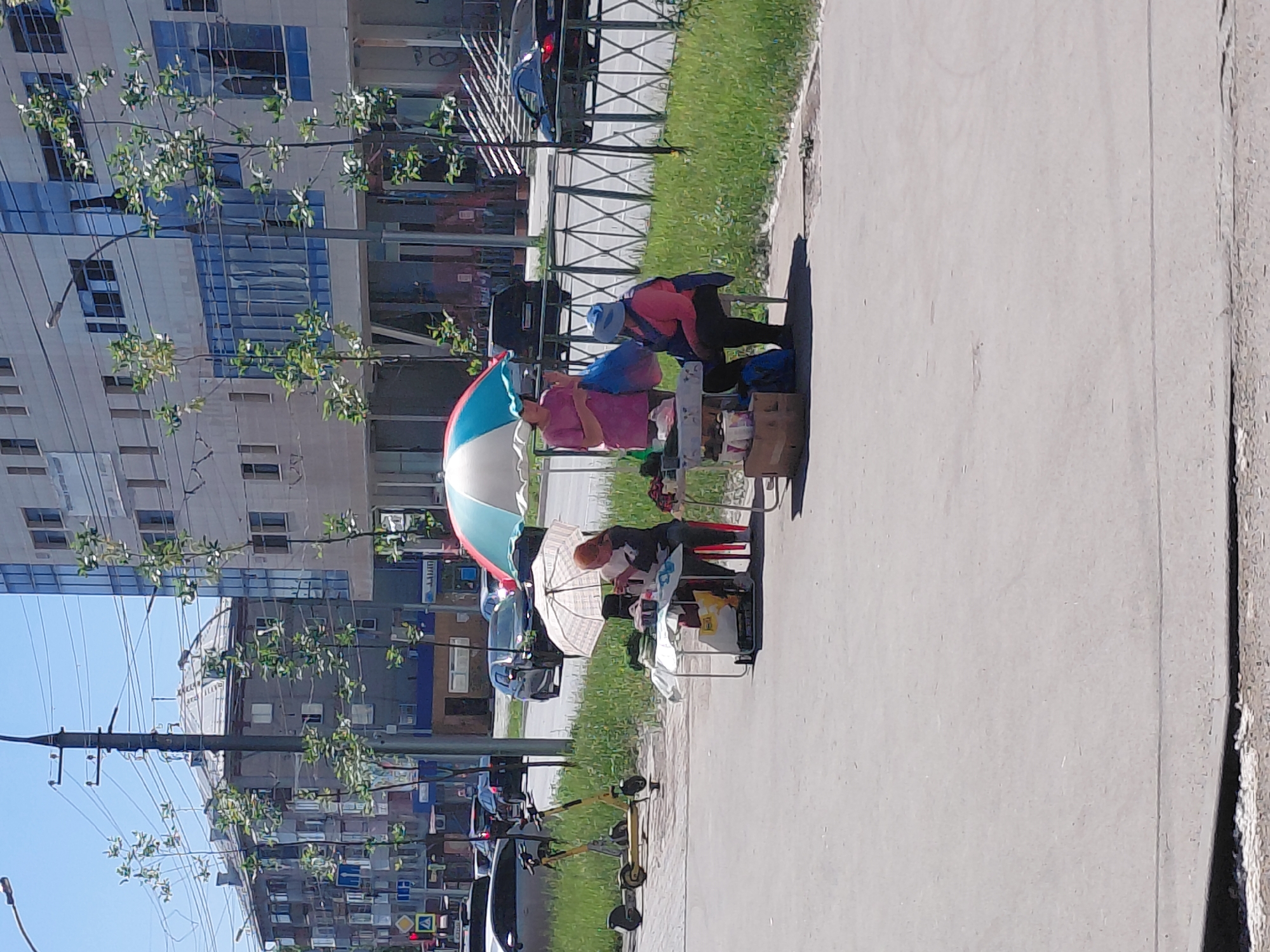 On a sunny day in an urban environment, two individuals are selling goods from a street stall. The stall is set up on a sidewalk next to a street with traffic. A multi-colored umbrella shades the area. One person, wearing a pink shirt, stands behind the table, holding a blue bag. Another person, wearing a blue head covering and a dark blue vest over a pink shirt, is seated at the table. Various items are displayed on the table and in boxes on the ground, suggesting they are merchandise for sale. In the background, there are buildings, trees, a utility pole with wires, and parked cars. A yellow electric scooter is visible on the sidewalk to the left of the stall. A metal fence runs along the edge of the sidewalk, separating it from a grassy area and the street. The scene appears to be a typical street market setup in a city.