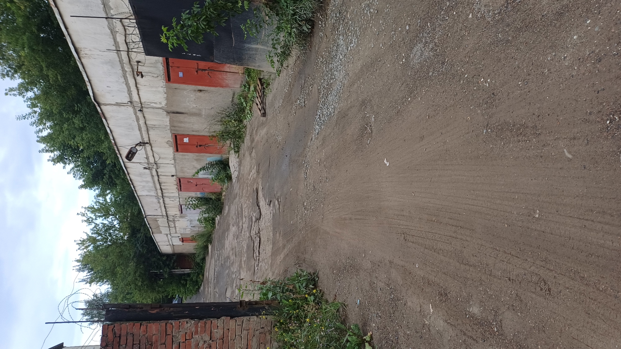 The image depicts a dirt road leading towards a row of garages. The garages are constructed of concrete with red metal doors. Some of the doors have visible locks or handles. Green vegetation grows around the garages and along the roadside, with some weeds present on the road itself. A brick wall and a metal gate are visible on the left side of the road, with barbed wire atop the gate. The sky is overcast. The scene appears to be in a somewhat neglected urban or industrial area.