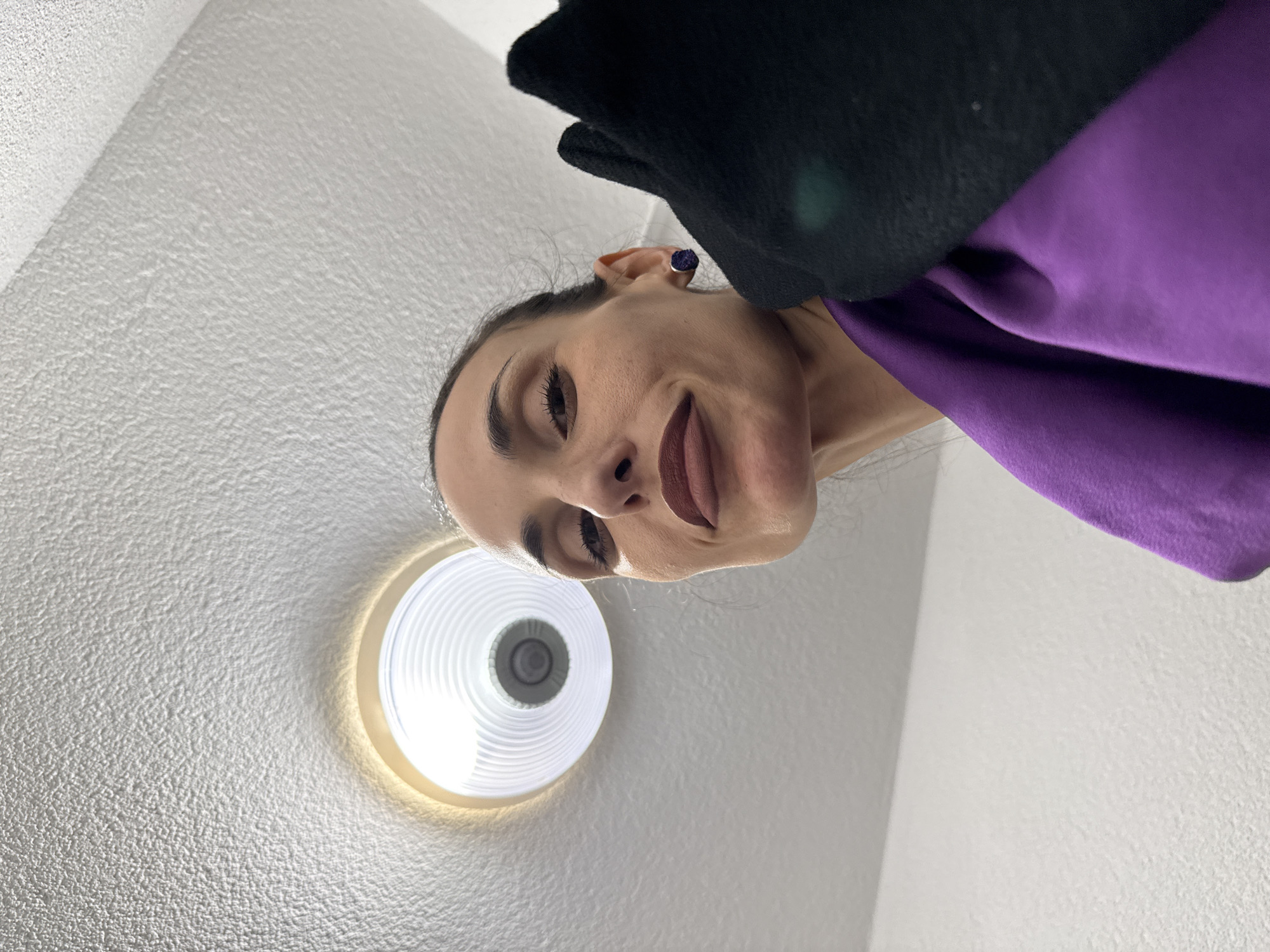 A woman is looking down at the camera, with a light fixture on the textured white ceiling visible above her. She is wearing a purple top and a black jacket. Her face is angled, and she has dark lipstick. The location in the city of Lovech, Bulgaria is not visually discernible from the image.