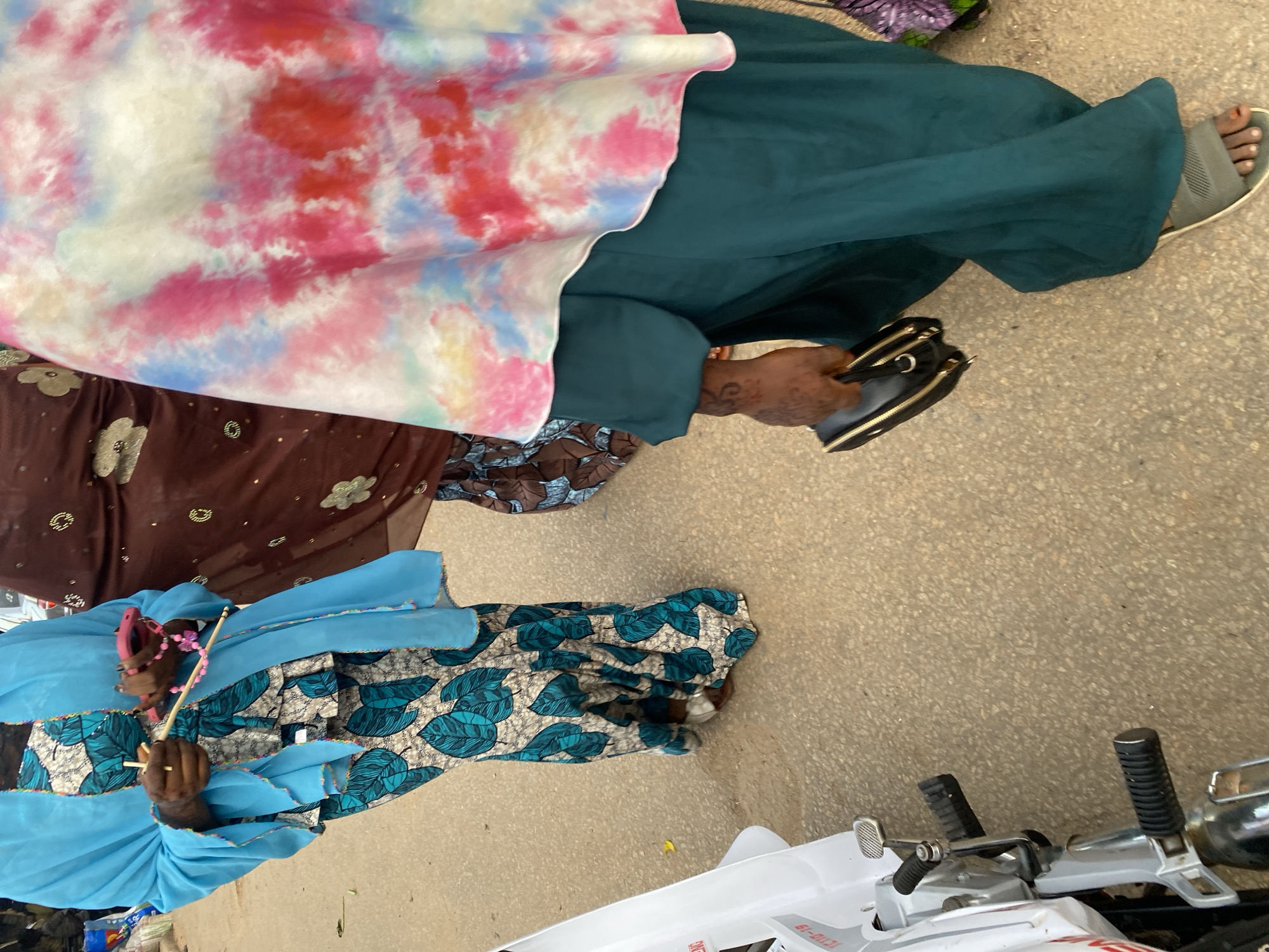 In Jos, Nigeria, three women are walking on a paved surface. The woman on the left wears a blue headscarf and a teal and white patterned dress with large leaf motifs. She is holding a pink object and two wooden sticks. The middle woman is wearing a brown garment adorned with gold floral embroidery. The woman on the right is draped in a tie-dye headscarf with pink, red, blue, and white patterns over a teal dress. She is carrying a black clutch purse. A white motorcycle is partially visible on the left side of the image. The ground is a textured beige concrete.
