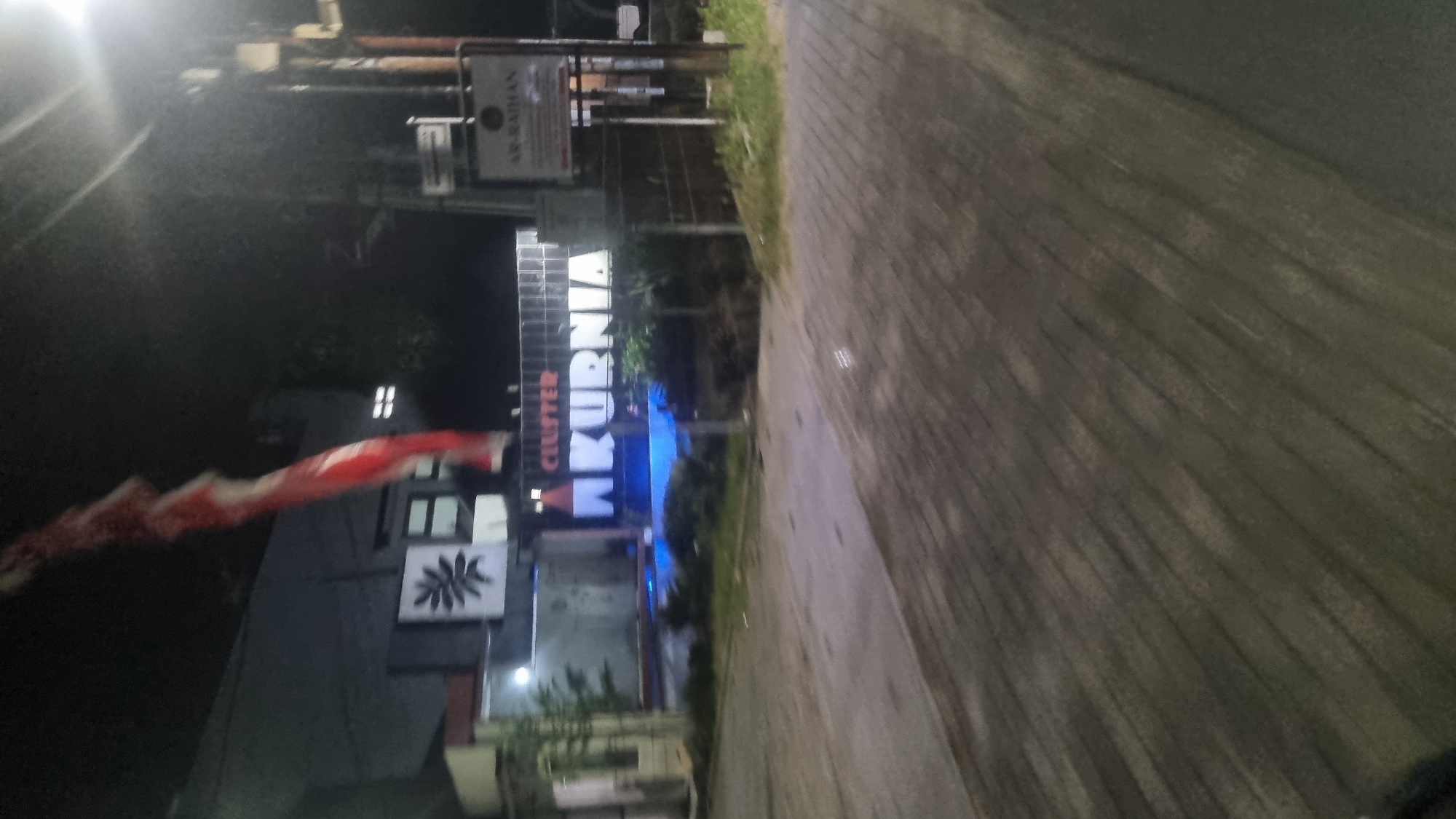 In the city of Diwek, Indonesia, at night, a street scene unfolds. A prominent sign, illuminated in bright white and accented with a blue glow from behind, reads "CLUSTER KURNIA". The sign is large and positioned centrally in the frame, suggesting it marks an entrance or a specific location within the cluster. To the right of this main sign, a smaller, rectangular sign is visible, with text that appears to read "AR-RAIHAN".
The foreground of the image is dominated by a paved road or walkway, rendered in shades of grey and brown due to the low light conditions and a subtle motion blur, indicating the photograph was taken while moving. The texture of the paving stones is discernible.
In the background, a building with dark walls and some illuminated windows can be seen. A red and white flag is displayed prominently, hanging vertically and partially obscuring the building. The flag's design suggests it might be the Indonesian flag, though the details are somewhat blurred. The overall environment is dark, indicative of nighttime, with artificial lighting from the signs and building windows providing the primary illumination. The scene appears to be an exterior view, possibly a residential area or a commercial complex.