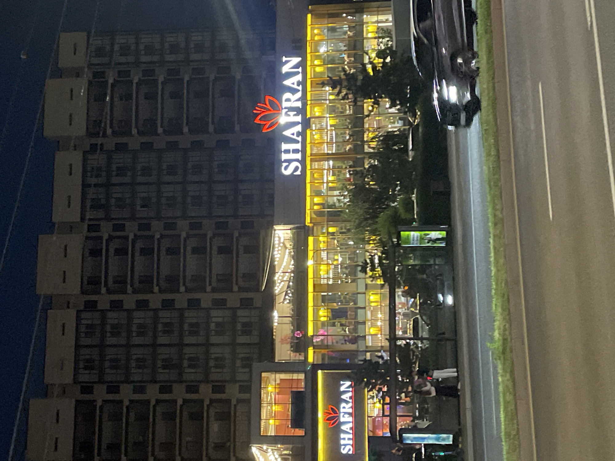 In Almaty, Kazakhstan, at night, a modern, multi-story building features a commercial establishment with illuminated signage. The signage displays "SHAFRAN" in white capital letters, accompanied by a red stylized floral logo. The commercial space has large glass windows revealing warmly lit interiors with patrons and decorative string lights. A dark-colored SUV is captured in motion on the right side of the image, driving on a road in front of the building. Two individuals are standing near the building on the left side, appearing to interact. The scene is set against a dark, twilight sky, suggesting an urban evening environment.