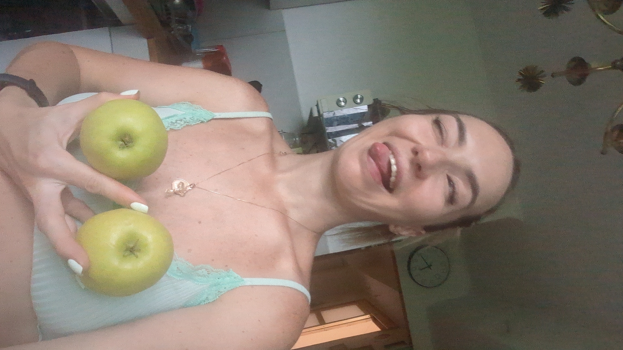 This media file shows a woman in a dimly lit indoor setting, possibly a kitchen. She is holding two green apples, one above the other, in front of her chest. She is wearing a light blue camisole with lace trim. Her right hand is positioned between the two apples, and her fingers are visible, as are her white-painted fingernails. A thin gold necklace with a small pendant is visible around her neck. The woman's face is tilted back, and she is sticking her tongue out, with her eyes closed or squinting, conveying a playful or mischievous emotion. The background is blurred, but suggests a domestic environment with some appliances and possibly a clock on the wall. There is no visible text in the image. The lighting appears to be artificial and suggests it could be nighttime or a dimly lit day.
