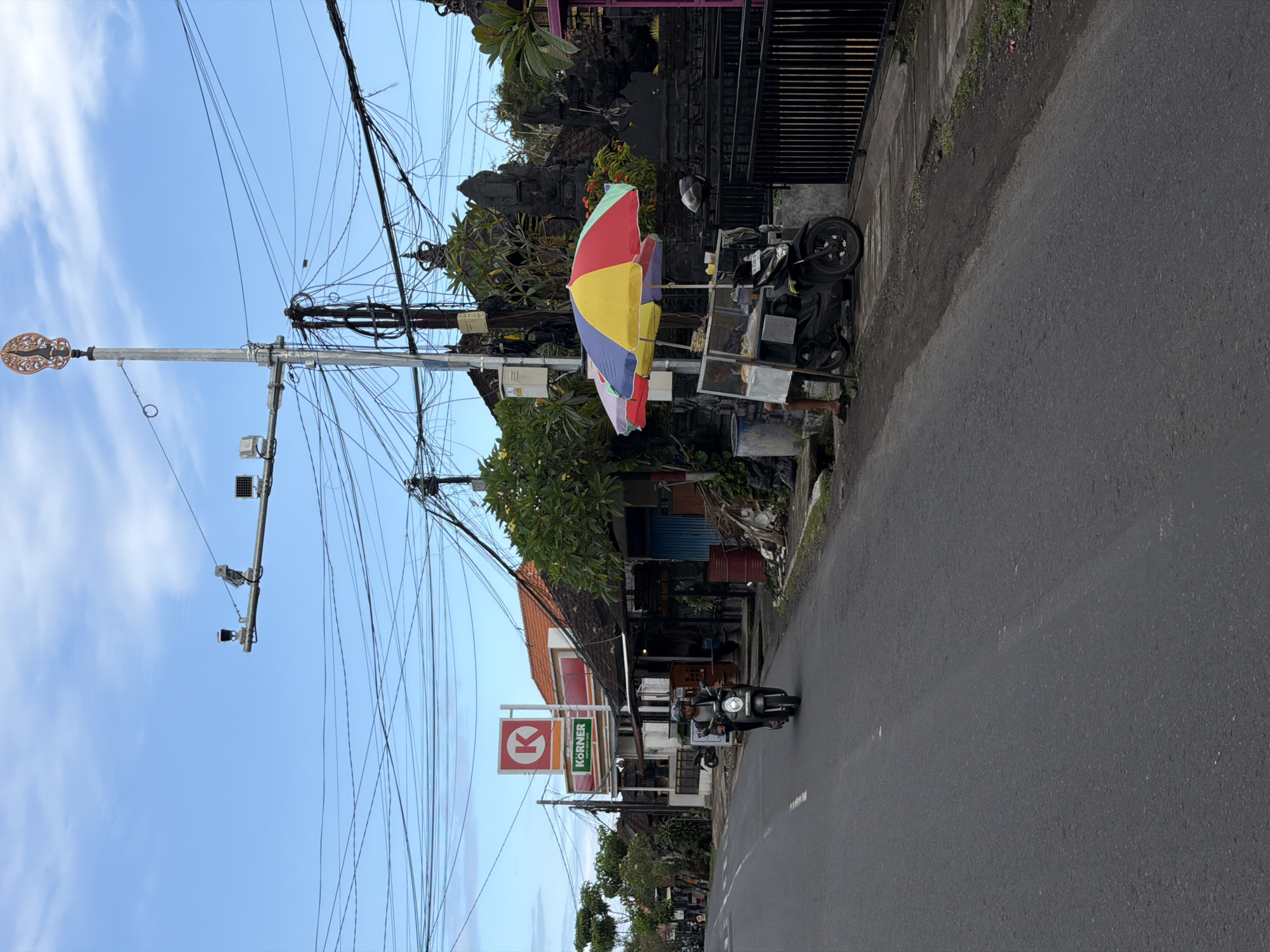 The photograph depicts a street scene in North Kuta, Indonesia. A black scooter with two riders travels on the right side of a paved road. Above them, a "KORNER" store sign is visible. Above the storefront, a tangle of electrical wires crosses the sky. A decorative street lamp stands to the left of the frame. On the right side of the road, near a property line, a colorful umbrella is erected next to a food vendor's cart. Vegetation and a dark fence are visible further down the road. The sky is blue with scattered clouds.