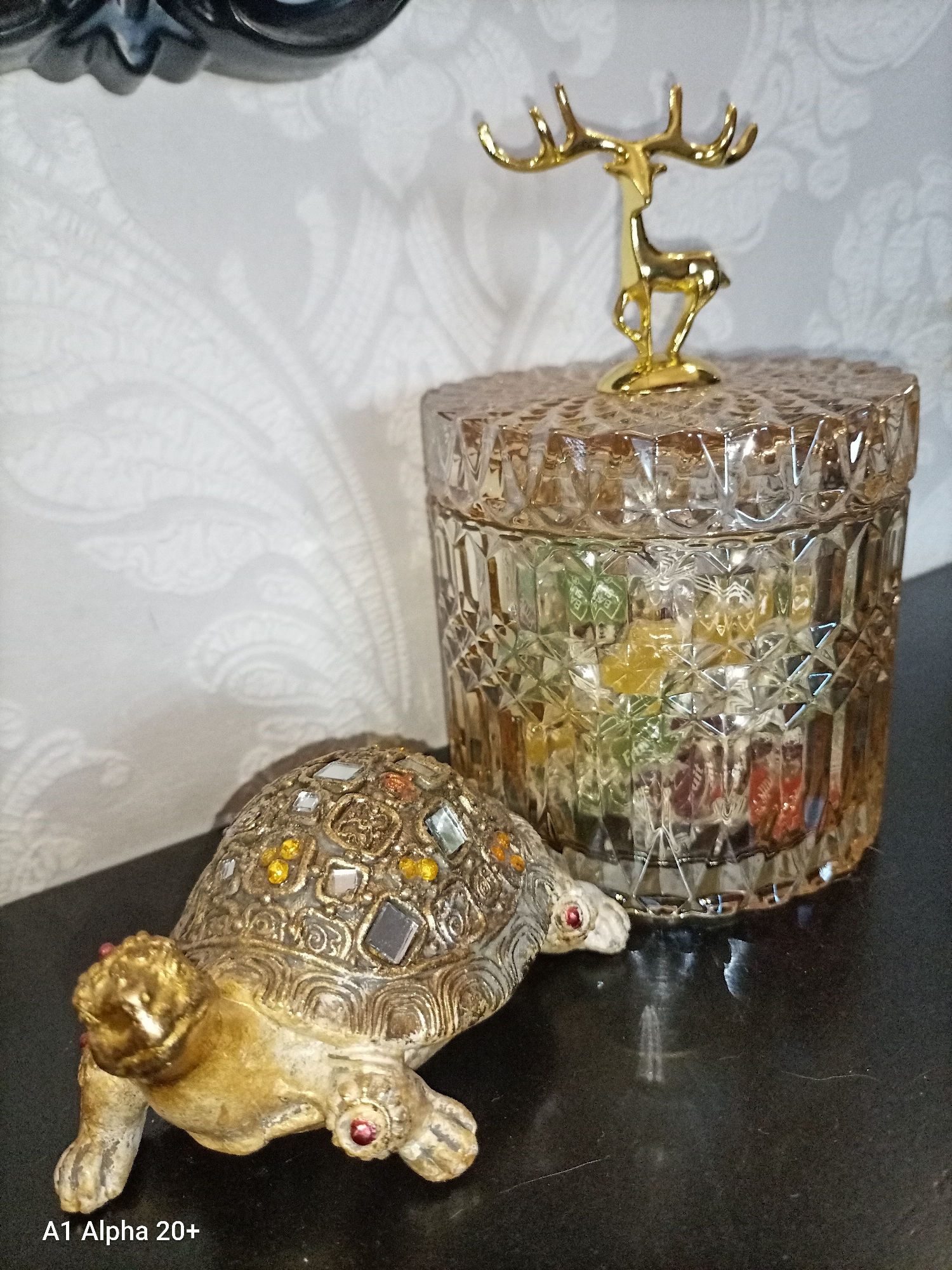 In a close-up shot with a shallow depth of field, a decorative tortoise figurine with a metallic gold and intricately patterned shell, embellished with colorful square and round gems, sits on a dark, reflective surface. To its right, a clear, faceted glass jar filled with colorful candies stands prominently. The lid of the jar is topped with a golden deer figurine, its antlers reaching upwards. The background features a textured wallpaper with a subtle white paisley pattern. The lighting suggests an indoor setting, casting soft shadows from the objects onto the dark surface and the wallpaper. The overall scene appears to be a still life arrangement of decorative items, possibly on a table or shelf. The location of Улукаўскі сельскі Савет, Belarus, is not discernible from the image.