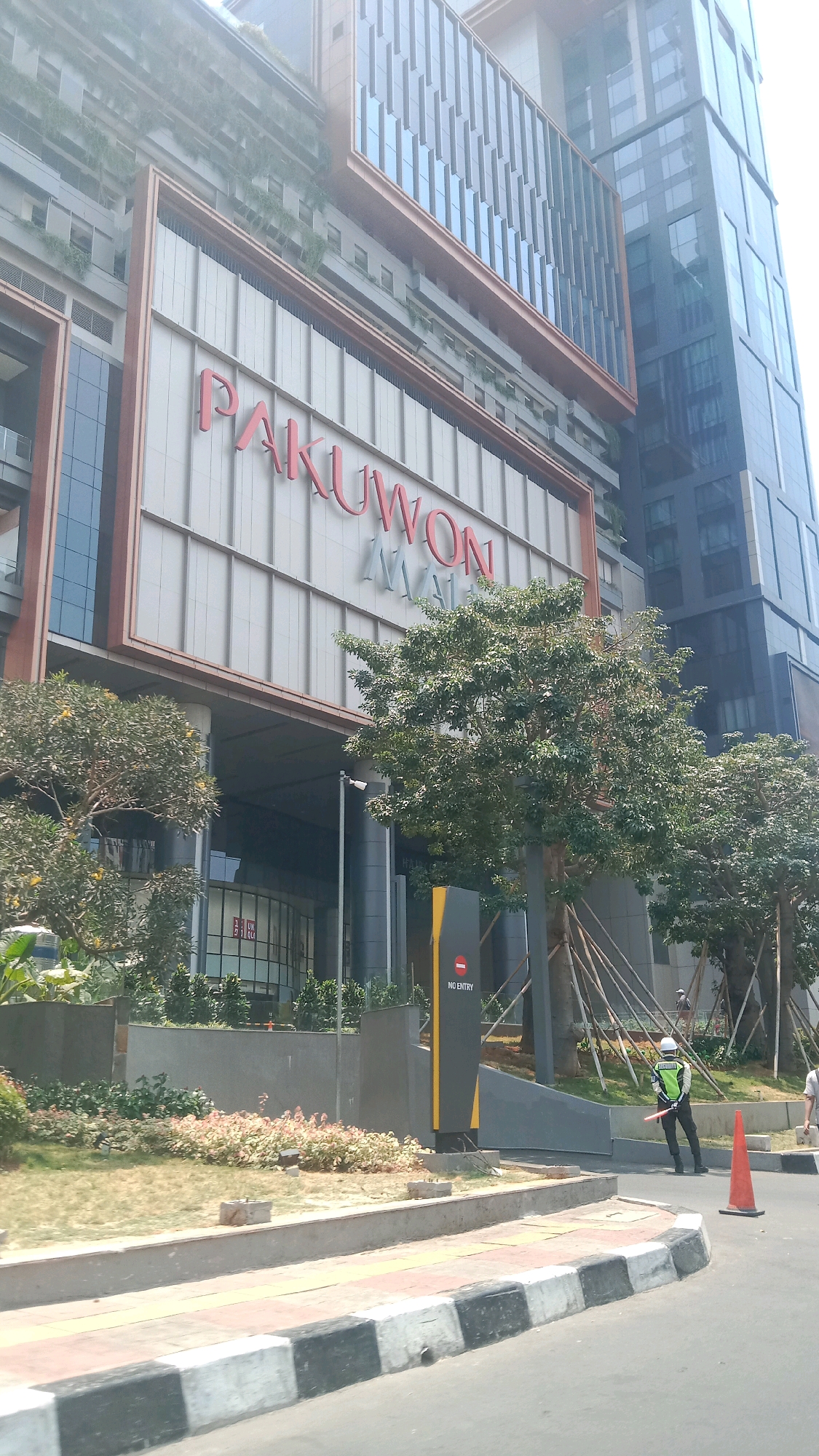 The image shows the facade of the Pakuwon Mall in Bekasi, Indonesia. The mall has a modern architectural design with large glass windows and a prominent sign with red lettering that reads "PAKUWON MALL". In front of the mall, there are trees and landscaping. A security guard wearing a reflective vest and a white helmet is standing near an entrance, holding a red stick. An orange traffic cone is placed on the road. The scene appears to be on a sunny day.
