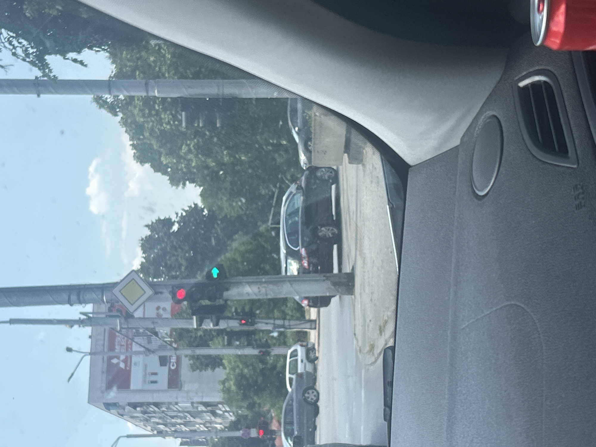 The image shows a street scene in Plovdiv, Bulgaria. In the foreground, the dashboard of a car is visible, with a red can of soda on the right side. The car appears to be stopped at a traffic light. In the middle ground, several cars are visible, including a dark-colored sedan and a white minivan. A traffic light is visible, showing a green light. In the background, a large building with a "Mitsubishi" advertisement is visible, along with trees and a blue sky with clouds. The traffic light is positioned at the intersection of Trakia Boulevard and Tsar Ivan Alexandar Street.