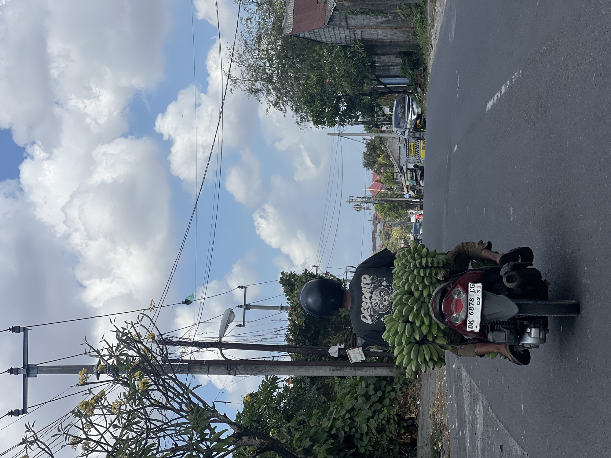 A person wearing a black helmet and a black t-shirt with the word "DESPAIR" is riding a red motorcycle loaded with large bunches of green bananas. The motorcycle is on an asphalt road with white lane markings, in what appears to be a residential or commercial area of Denpasar, Indonesia. The person is facing away from the camera, looking down the road. The environment includes trees, power lines with cables, a utility pole, a building with a corrugated metal roof, and other vehicles in the distance. The sky is blue with scattered white clouds. The license plate on the motorcycle is visible and reads "DK 6878 CG".