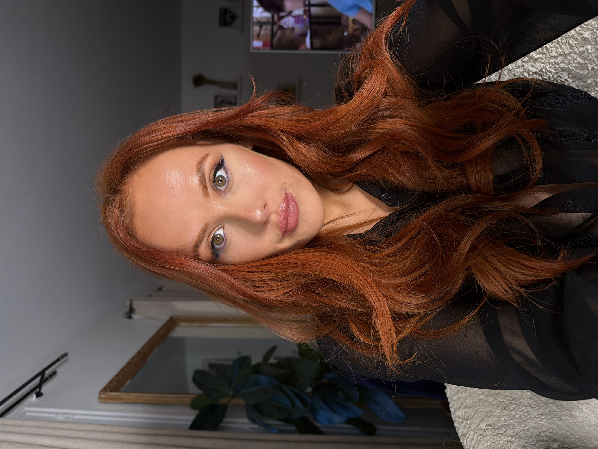 A person with red hair is lying on a surface, wearing a black sheer top. The background includes a partially visible plant, a mirror, and a television screen. The location is not identifiable as Paris, France.