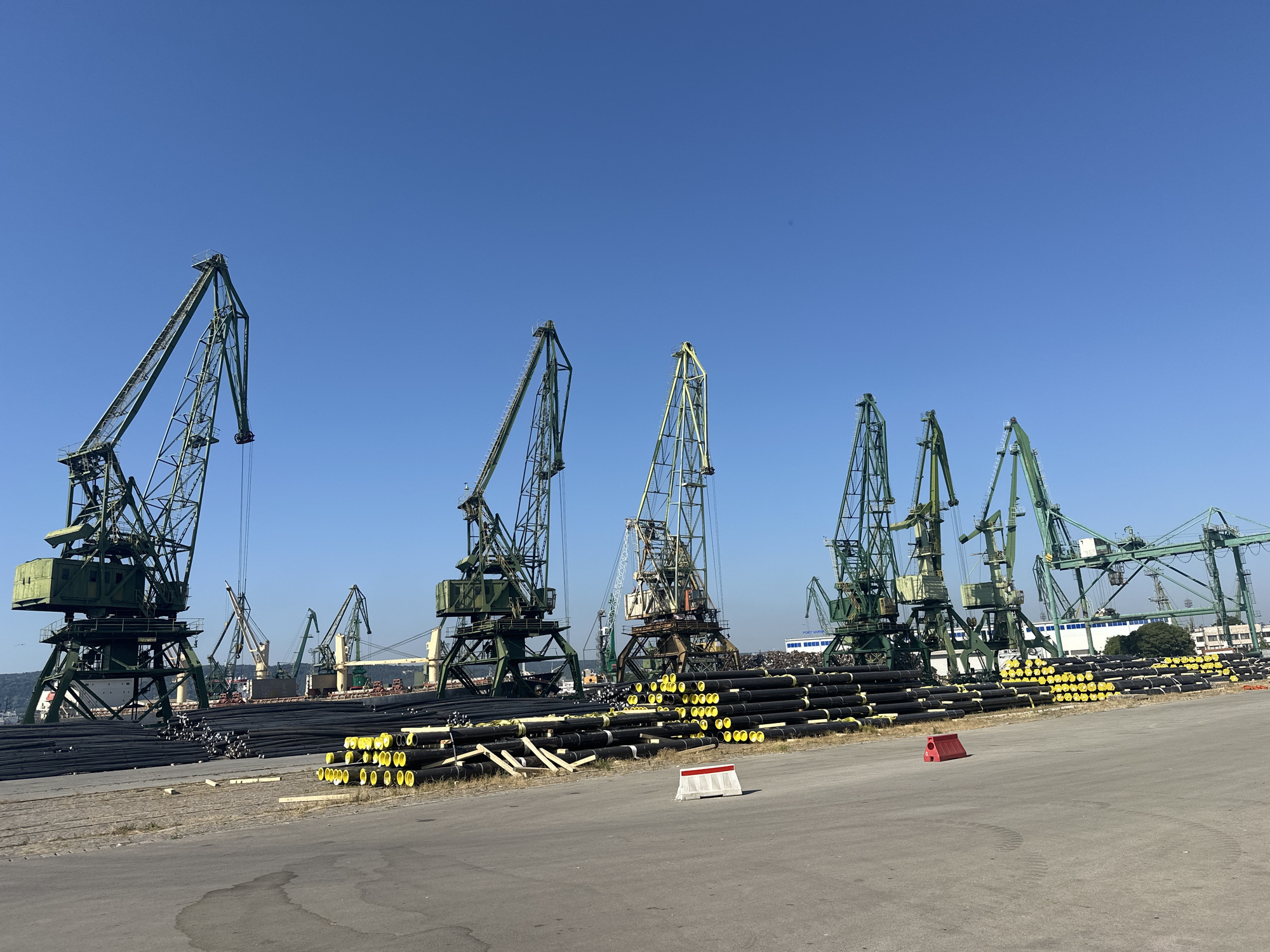 A wide shot captures a harbor scene under a clear blue sky. A row of tall, green industrial cranes dominates the frame, their massive structures silhouetted against the sky. In the foreground, neatly stacked bundles of black pipes with yellow markings lie on the concrete ground. Beyond the pipes, a large cargo ship is visible, partially obscured by the cranes. The environment suggests a busy port or industrial area, possibly involved in shipping or manufacturing.