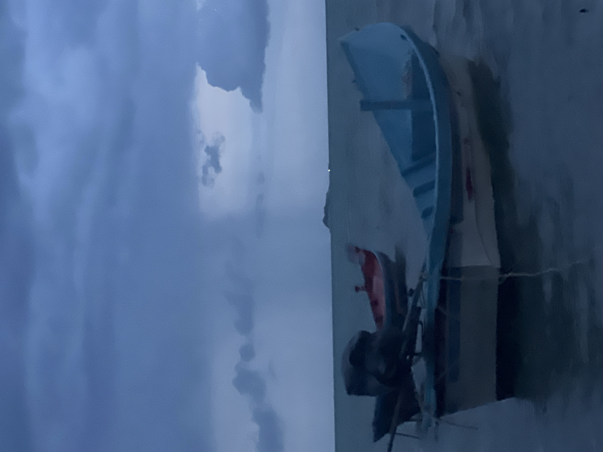The image depicts a serene, twilight scene at sea, likely near a coastal area, with several small boats bobbing gently on the water. The sky is overcast with heavy, dark blue and grey clouds, suggesting an impending storm or the aftermath of one. A faint silhouette of a landmass or island is visible on the horizon. In the foreground, a light blue and white boat is prominently featured, its hull partially submerged in the dark, rippling water. Behind it, another boat, colored red and white, is anchored. A third boat, mostly obscured by darkness, is visible to the left, with an outboard motor mounted on it. The overall ambiance is somber and tranquil, dominated by muted blues and greys.