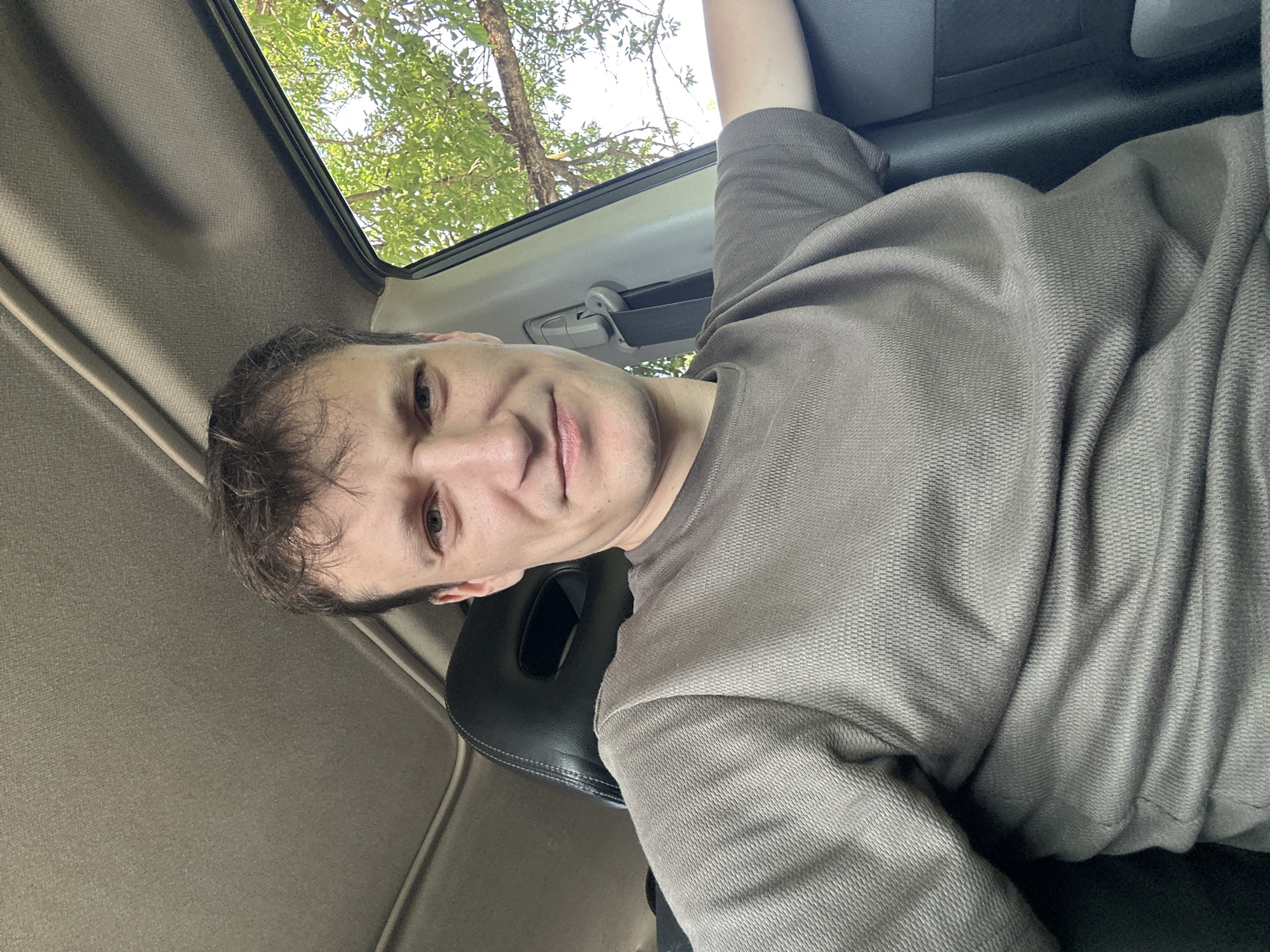 A young man with light brown hair and a light complexion is sitting in the driver's seat of a car, looking at the camera. He is wearing a taupe-colored t-shirt. The interior of the car is visible, including the black leather headrest of the seat behind him, the car's ceiling, and a part of the car door with a seatbelt. Through the open car window, green trees and blue sky are visible. The scene appears to be outdoors, in a natural setting. There is no information in the image to suggest the location is in the city of Makiv, Ukraine.