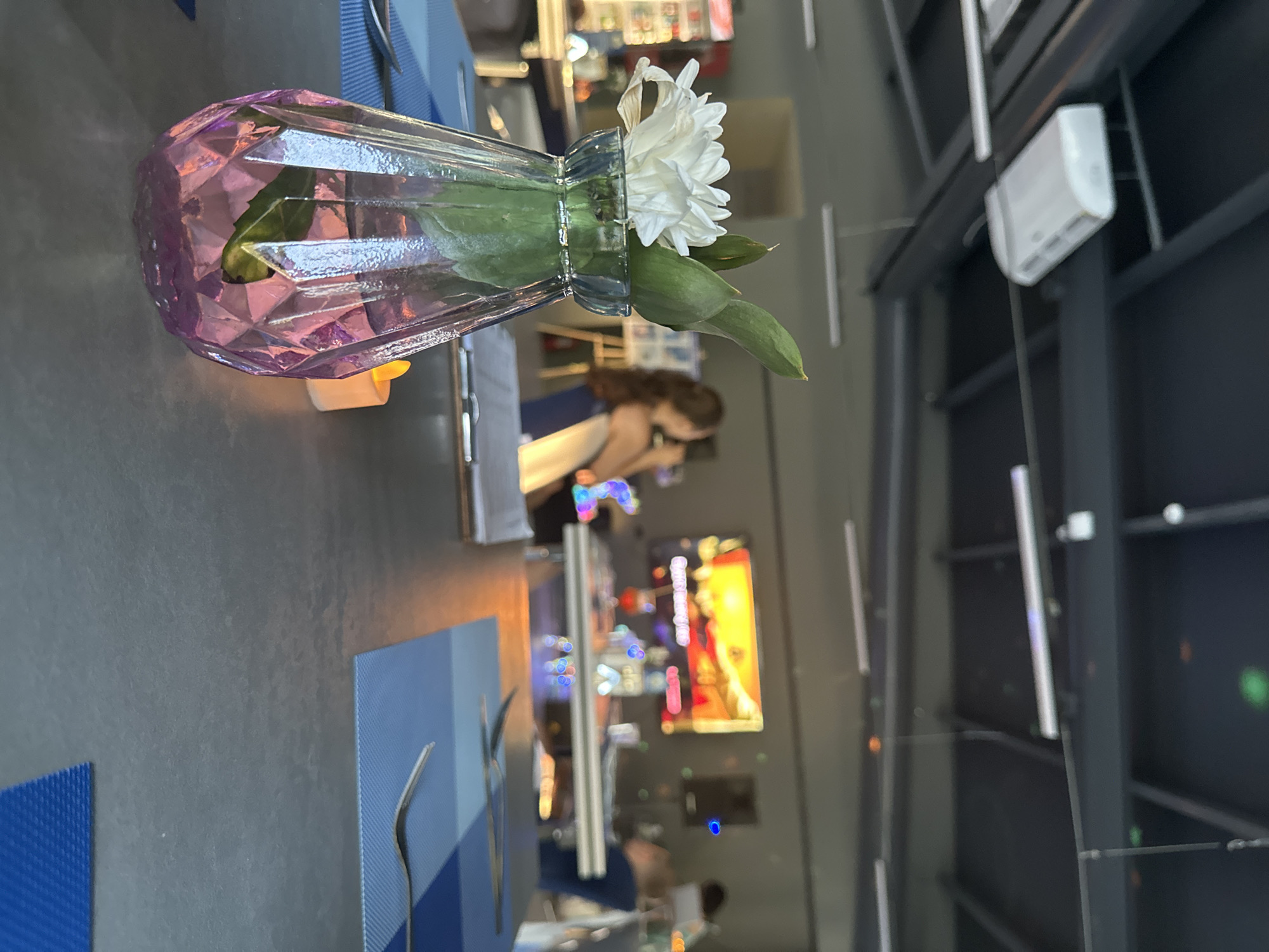 The image shows a close-up of a decorative glass vase on a table, with a single white flower and green leaves inside. The vase has a faceted design and a pinkish hue. A small white candle is visible behind the vase, emitting a soft glow. The table is covered with a dark grey surface, and in the foreground, there are blue placemats and cutlery. In the background, there are out-of-focus elements of a restaurant or bar setting, including people, screens displaying colorful content, and industrial-style ceiling structures with lighting. One person is visible in the middle ground, facing away from the camera, with long brown hair. The overall impression is that of a dimly lit, modern interior, possibly during an evening gathering or meal. The location context indicates Danilovka, Altai Krai, Russia.