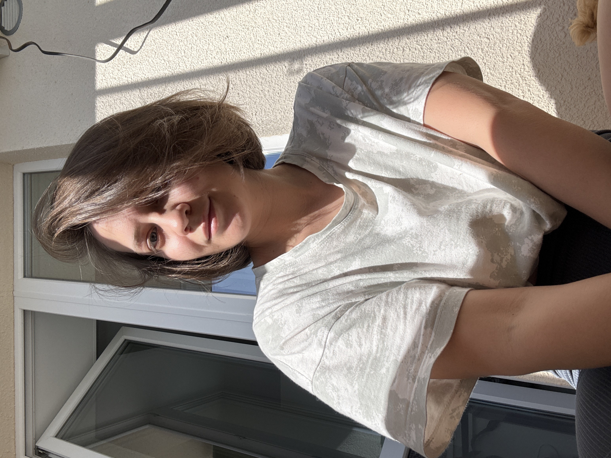 A woman with short brown hair is looking at the camera, possibly on a balcony. She is wearing a patterned white t-shirt and dark pants. There is a window with an open pane behind her. The background features a textured wall and a glimpse of an electrical cord. The photo appears to be taken in daylight. The location in the city of Pervomaysky, Russia, is unverifiable from the provided image.