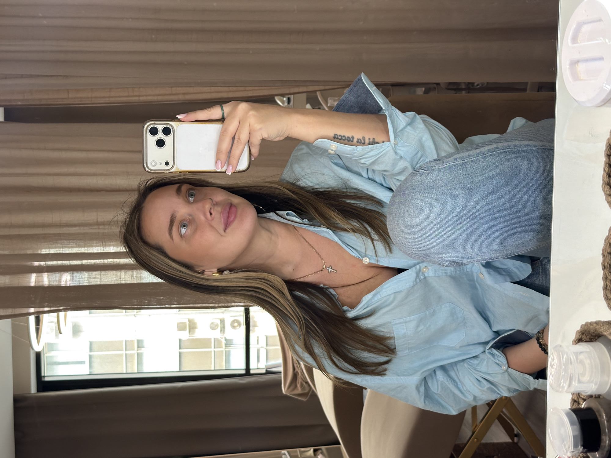 A woman is taking a selfie in what appears to be an indoor setting, possibly an apartment or hotel room. She is holding a smartphone, specifically an iPhone, in her right hand, capturing her image. The woman is wearing a light blue button-down shirt and jeans. She has light brown hair with blonde highlights and a necklace with a cross pendant. Her left arm has a tattoo.
In the background, there is a window with multiple panes and what seems to be air conditioning units installed. The presence of multiple residential or commercial buildings is evident through the window, suggesting an urban environment. Given the architectural style and the general appearance of the buildings, it's plausible that the location could be in Moscow, Russia. There are brown curtains or blinds partially obscuring the window. A make-up mirror or vanity with various cosmetic items on it is visible on the right. There is a chair and a beige wall in the background.